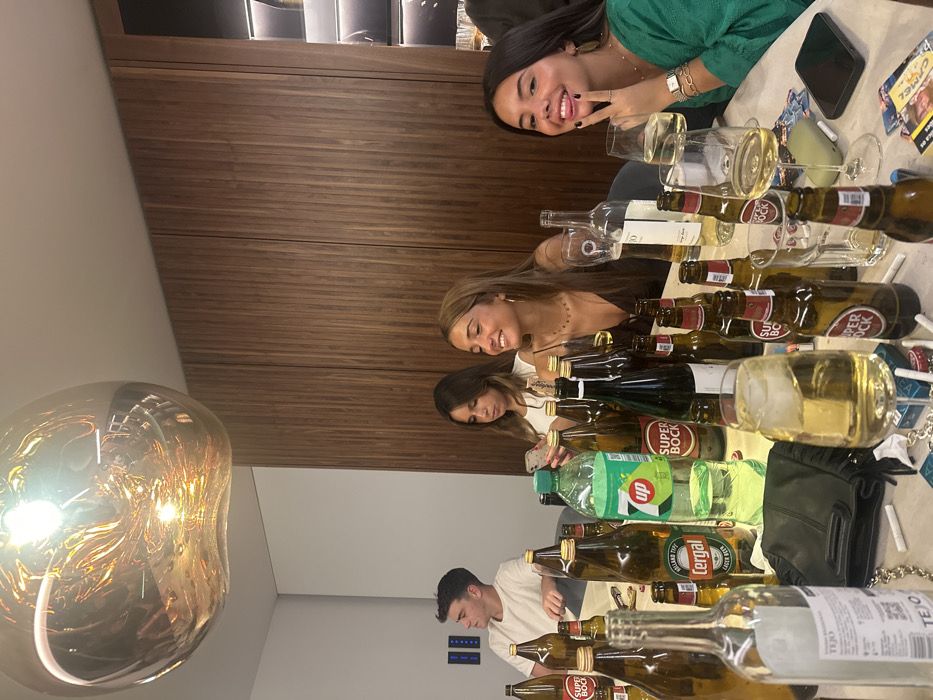 The image captures an indoor social gathering, ostensibly located within Lordelo do Ouro e Massarelos, Portugal, around a light-colored table. Four young individuals are seated. On the far left, a male with short dark hair, wearing a white t-shirt, looks towards the left side of the frame. To his right, a female with long dark hair, wearing a white top, is focused on a smartphone held in her hands. Adjacent to her, a second female with long light brown hair, in a dark V-neck top, smiles towards the right. On the far right, a third female with long dark hair, wearing a green top, looks directly at the camera with her tongue slightly extended and makes a peace sign gesture with her right hand.
The table surface is cluttered with numerous glass bottles, predominantly brown beer bottles labeled "SUPER BOCK" and "Coral Cerveja Especial," alongside a clear glass bottle labeled "TEJO," and a green 7-Up soda bottle. Multiple empty and partially filled glasses, some containing a light-colored liquid, are dispersed across the table. Also present are a black bag, several loose cigarettes, cigarette butts, and mobile phones.
The environment includes a white ceiling from which a large, illuminated, amber-tinted spherical light fixture hangs centrally in the foreground. The rear wall features vertical dark wood paneling, while the wall to the left is a plain light color. A partially visible, internally lit shelving unit is present on the far right, adjacent to the wooden paneling.
