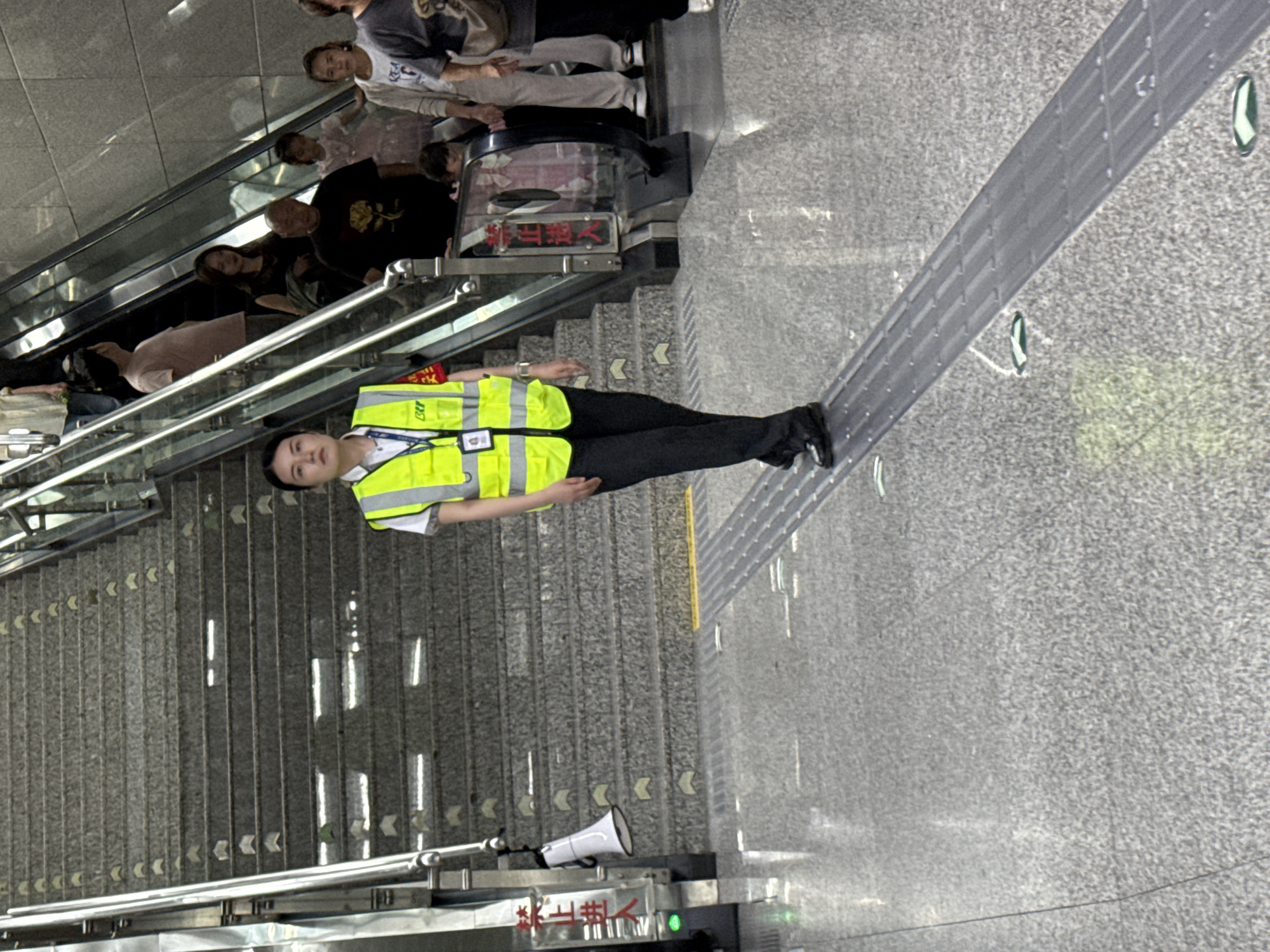 An indoor scene in what appears to be a metro station or public transport hub in Yuzhong District, China, is depicted. The image has been rotated 90 degrees clockwise from its original orientation.
In the foreground, a staff member is standing, facing slightly towards the viewer. They are wearing a white short-sleeved shirt, dark trousers, and a bright yellow high-visibility vest with reflective stripes and a badge on a lanyard, which appears to have "CPY" or "CST" written on it. Their expression is neutral and alert.
To the left of the staff member, an escalator is visible, moving people upwards. Several individuals are on the escalator, including a woman with long dark hair, a man with a bald head and a dark t-shirt with a yellow design, and other indistinct figures. Beside the escalator, a staircase with gray steps and metal handrails descends. Many steps have white chevron markings indicating direction.
The floor is a light gray, textured surface, possibly granite or concrete. A raised, gray tactile paving strip runs across the floor in front of the staff member, likely for the visually impaired.
Near the base of the staircase/escalator, a white megaphone is placed on a railing. Below it, a red sign with white characters clearly states "禁止逆行" (Jìnzhǐ Nìxíng), meaning "No counter-flow" or "Do not go against the flow". Another sign, less distinct but visible above the escalator's entrance/exit, reads "禁止带犬" (Jìnzhǐ Dài Quǎn), which translates to "No dogs allowed" or "Pets are forbidden."
The lighting suggests an indoor setting, likely well-lit by artificial means, or during the daytime. The overall impression is one of a busy public space with clear safety regulations.