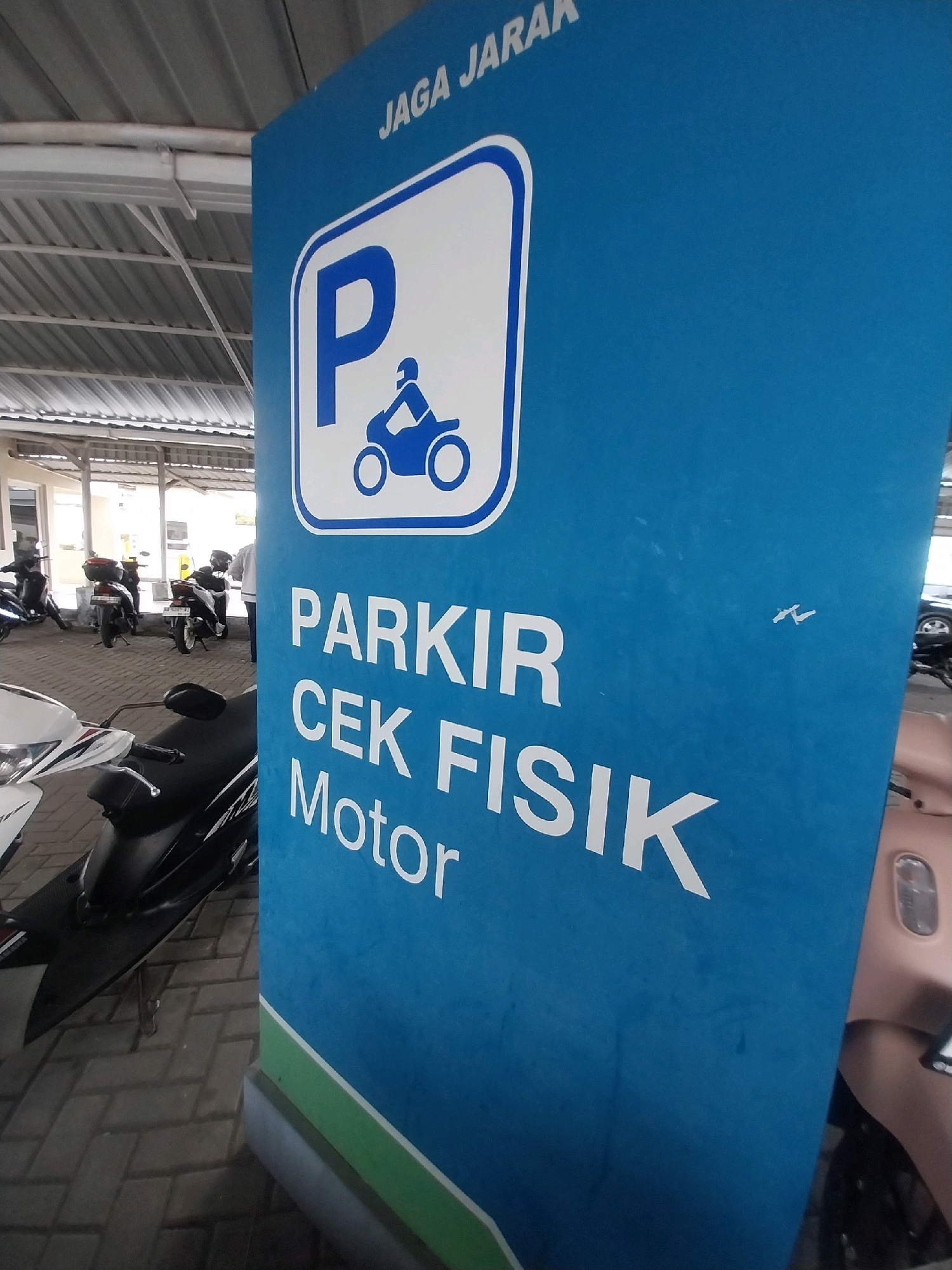 A blue sign with white text and a motorcycle parking symbol is prominently displayed in the foreground. The sign reads "JAGA JARAK" at the top, followed by a circular white symbol with a blue outline containing a blue silhouette of a motorcycle and rider. Below this, the text "PARKIR CEK FISIK Motor" is written in large white letters. The sign is positioned in what appears to be an outdoor parking area, possibly at a public facility in Sleman, Indonesia, given the text language and common parking regulations in the region. The background shows a covered parking area with a metal roof structure, tiled flooring, and several parked motorcycles and scooters. The lighting suggests it is daytime.