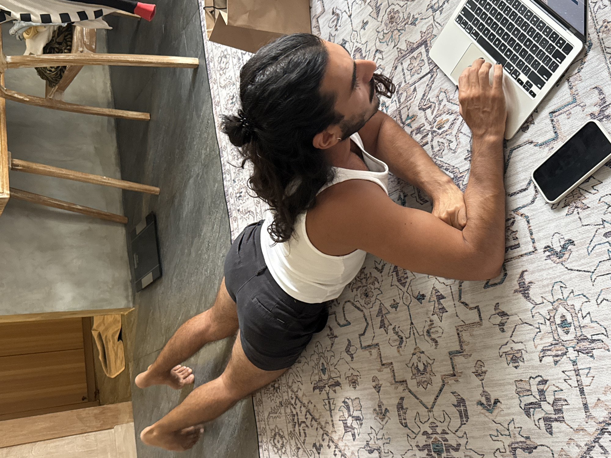 A man with long, dark, curly hair tied in a bun is lying on his stomach on a patterned rug in what appears to be an indoor residential setting. He is wearing a white tank top and dark shorts. The man is looking down at a laptop that is open in front of him, with his hands on the keyboard and trackpad. Next to the laptop is a smartphone. In the background, a wooden table and chair are visible, along with a brown paper bag. The floor is tiled, and beyond that, a wooden doorframe leads to another area with a tan blanket on the floor. A digital scale is also visible on the tiled floor. This scene is located in the city of North Kuta, Indonesia.