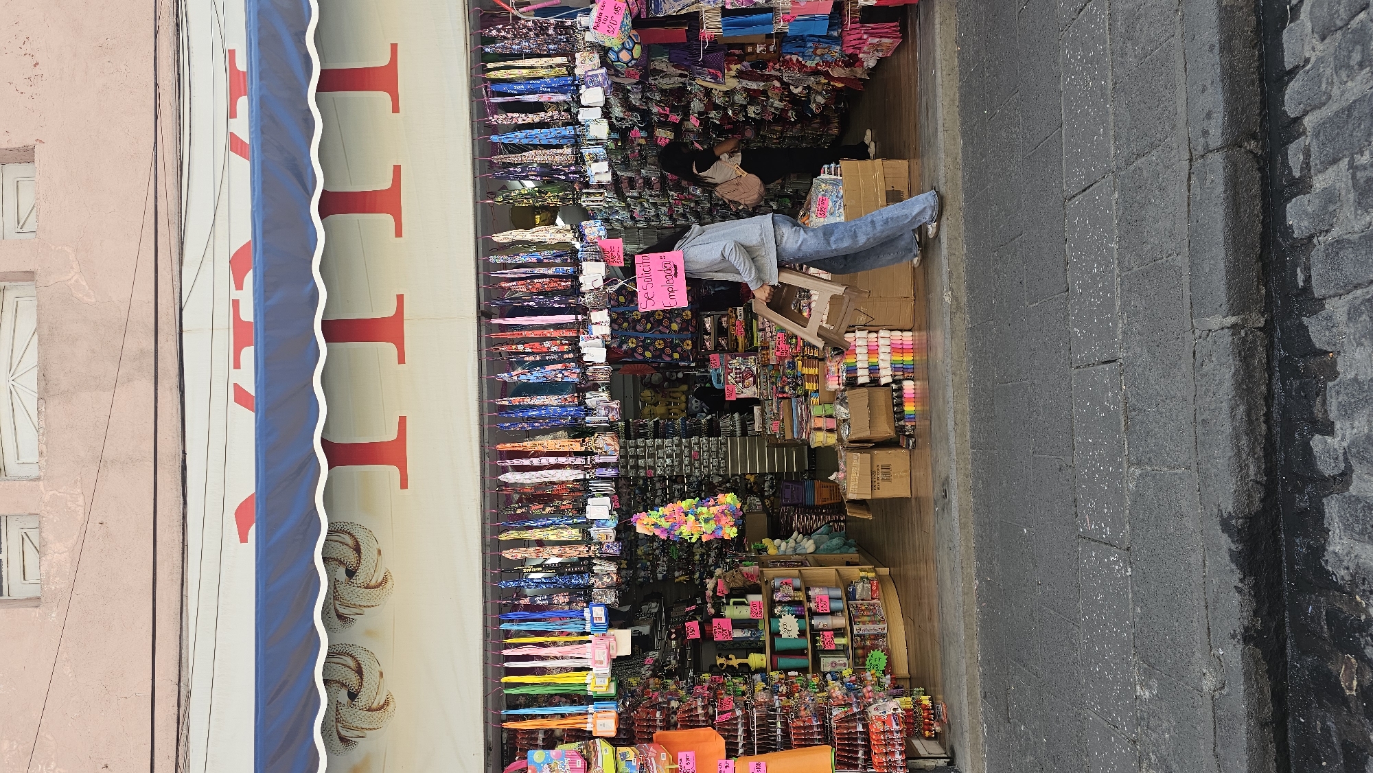This image captures a vibrant street scene outside a bustling storefront, likely a general store or novelty shop, in Puebla, Mexico. The perspective is looking into the shop from the street level.
The shop entrance is packed with an extensive display of colorful merchandise, spilling out towards the sidewalk. Items are hung on multiple vertical racks and arranged on shelves and counters. These appear to include various accessories, small toys, craft supplies like spools of ribbon, and numerous other colorful small goods. Among the prominent items are rows of patterned umbrellas or parasols.
A person, wearing a light-colored hoodie and blue jeans, stands just inside the shop entrance, holding a small, light brown plastic stool. Attached to their chest or hanging from their neck is a pink sign that clearly reads: "Se solicita empleado!" (Employee wanted!). Further inside and behind a counter, another person in dark clothing is partially visible, likely an employee attending to the shop.
Above the storefront, a blue and white striped awning is extended. Red letters are visible on the white part of the awning, possibly forming a shop name. On the left side of the image, the facade of a light pink building with white-framed windows is visible, suggesting typical urban architecture.
The street or sidewalk in the foreground is paved with dark gray blocks or cobblestones, and there's a darker, possibly damp patch, closer to the right edge. Small pink and green price tags are scattered among the merchandise, some showing prices like "$2/10" and "$5". The overall impression is that of a lively commercial area during daylight hours.