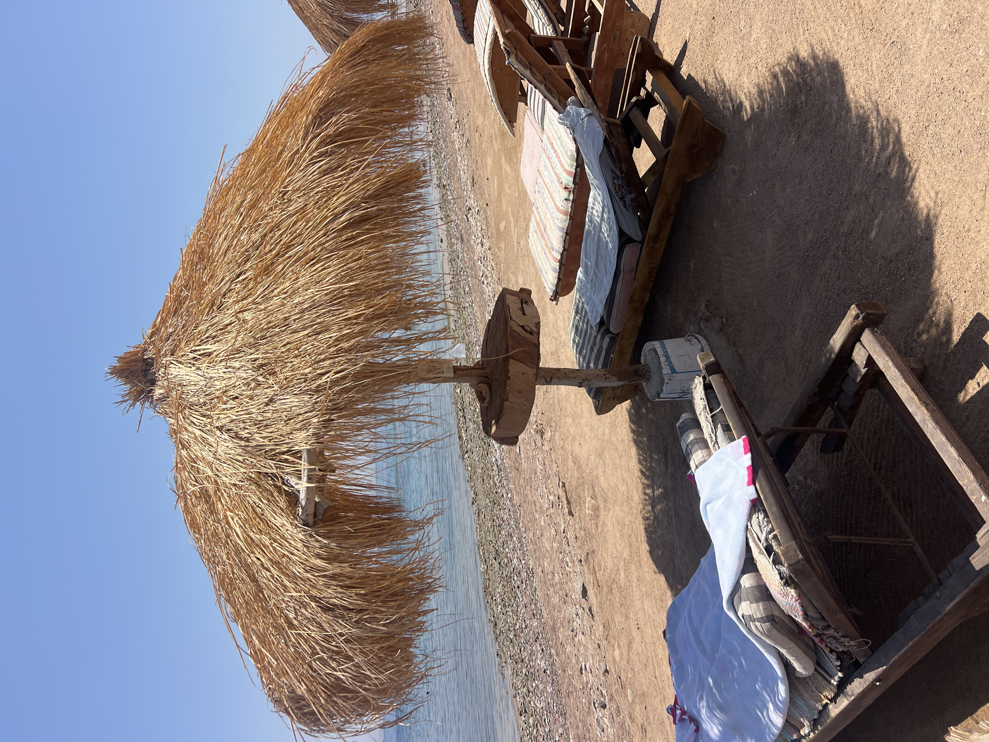 A straw umbrella shades two empty lounge chairs on a sandy beach next to a body of water. The beach is made up of sand and small pebbles. The water is calm and appears to be a light blue. The sky is clear and blue. The lounge chairs are made of wood and have cushions on them. There are towels on one of the lounge chairs. The scene is peaceful and relaxing. The location is Unknown, Egypt.