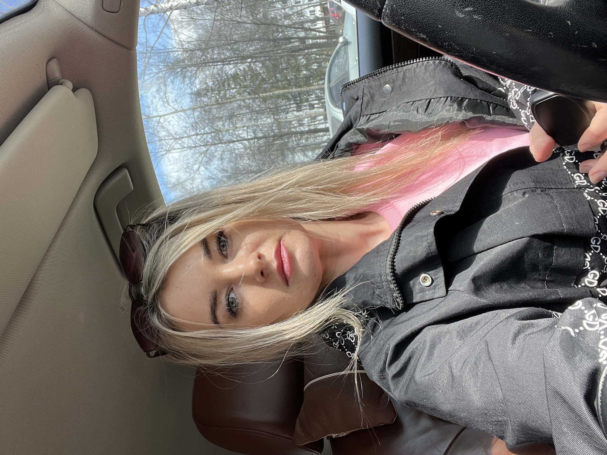A young woman is featured in a selfie taken inside a car in Chaikovsky, Russia. She has long, straight blonde hair, light-colored eyes, and rosy lips, looking directly at the camera with a neutral expression. Sunglasses are perched on her head. She is wearing a black collared jacket over a pink long-sleeved top. A black and white patterned fabric, possibly a bandana or jacket lining, with repeated "GD" text, is visible around her neck/collar area.
The setting is a car interior, illuminated by strong natural sunlight that creates distinct shadows. Visible car components include a light brown/beige headliner, a sun visor, and a dark car seat headrest. Part of a dark steering wheel or a passenger-side grab handle is visible in the upper right. An "AIRBAG" label is clearly printed on the car's ceiling panel. Through the car windows, numerous bare trees against a bright, clear sky indicate a sunny late winter or early spring day. The overall scene suggests a casual moment during travel.
