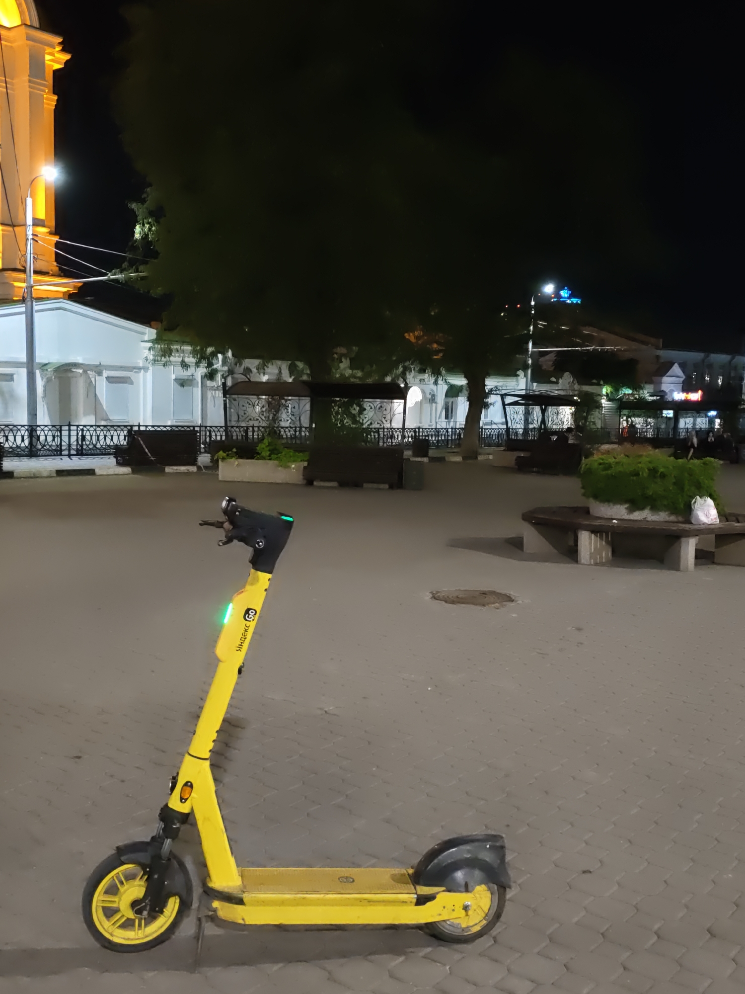 A yellow electric scooter, branded with "Yandex Go", is parked on a paved walkway at night. The scooter is positioned diagonally, with its handlebars facing left and its rear wheel slightly towards the camera. A green light is visible on the scooter's handlebars, and another green light is on the stem. In the background, there is a large tree with dark foliage, a white building with arched windows and ornate railings, and other structures that suggest an urban environment. Benches are visible in the mid-ground, and a rounded bench with a planter is in the right mid-ground. The scene is illuminated by artificial lighting, casting shadows. The sky is dark, indicating nighttime. The location is identified as the city of Rostov-on-Don, Russia.