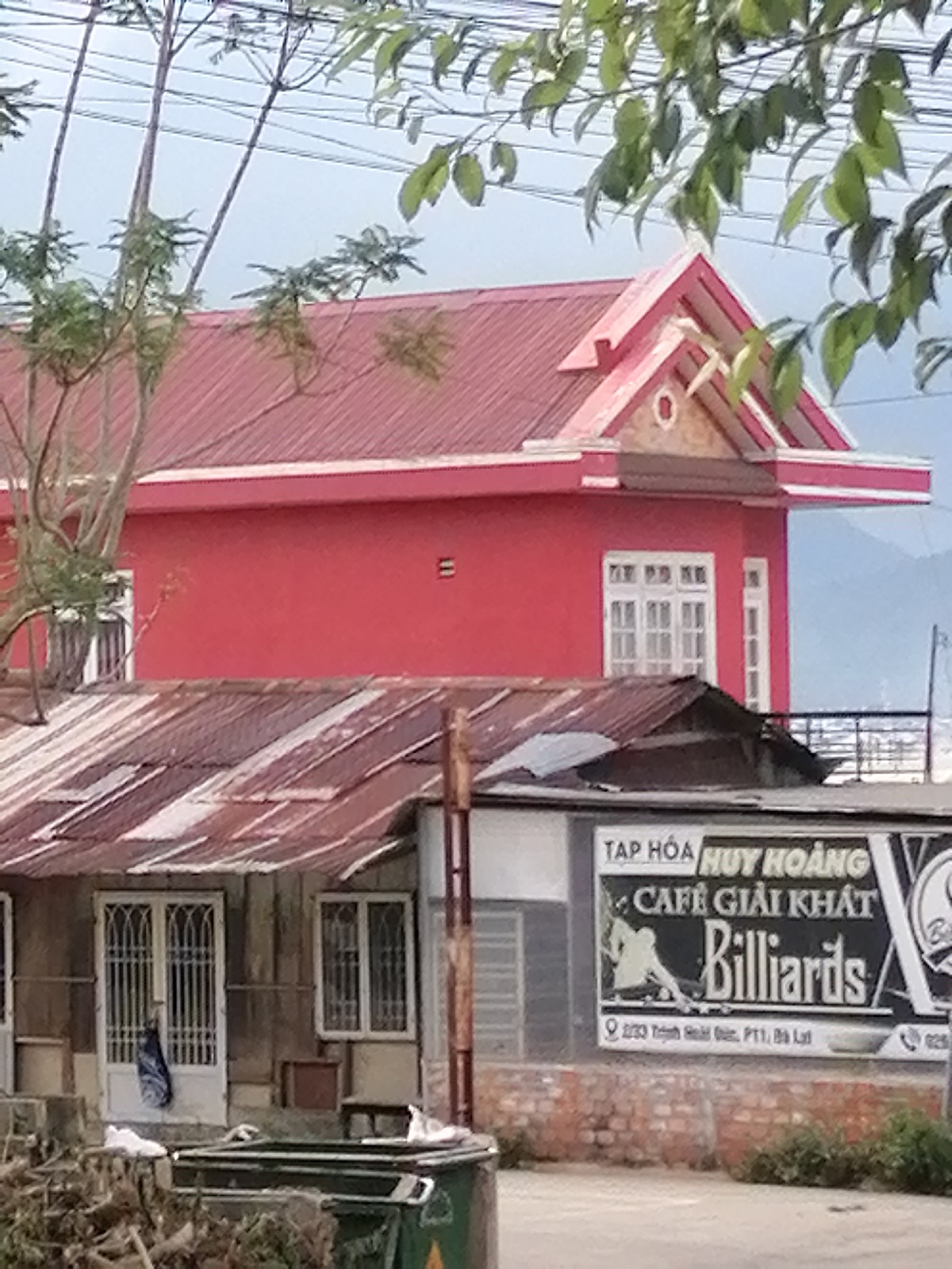 The image shows a street scene in Xuân Trường - Đà Lạt, Vietnam. In the foreground, a green dumpster is overflowing with debris. Behind it, a building with a corrugated metal roof is visible, with two white doors and windows featuring decorative metal grates. A blue bag hangs on one of the doors. Further back, a bright red building with white trim and multiple windows stands prominently. Above the red building, trees with green leaves and branches extend into the frame. A sign on a lower building reads "TẠP HÓA HUY HOÀNG CAFE GIẢI KHÁT Billiards". The sky is overcast.