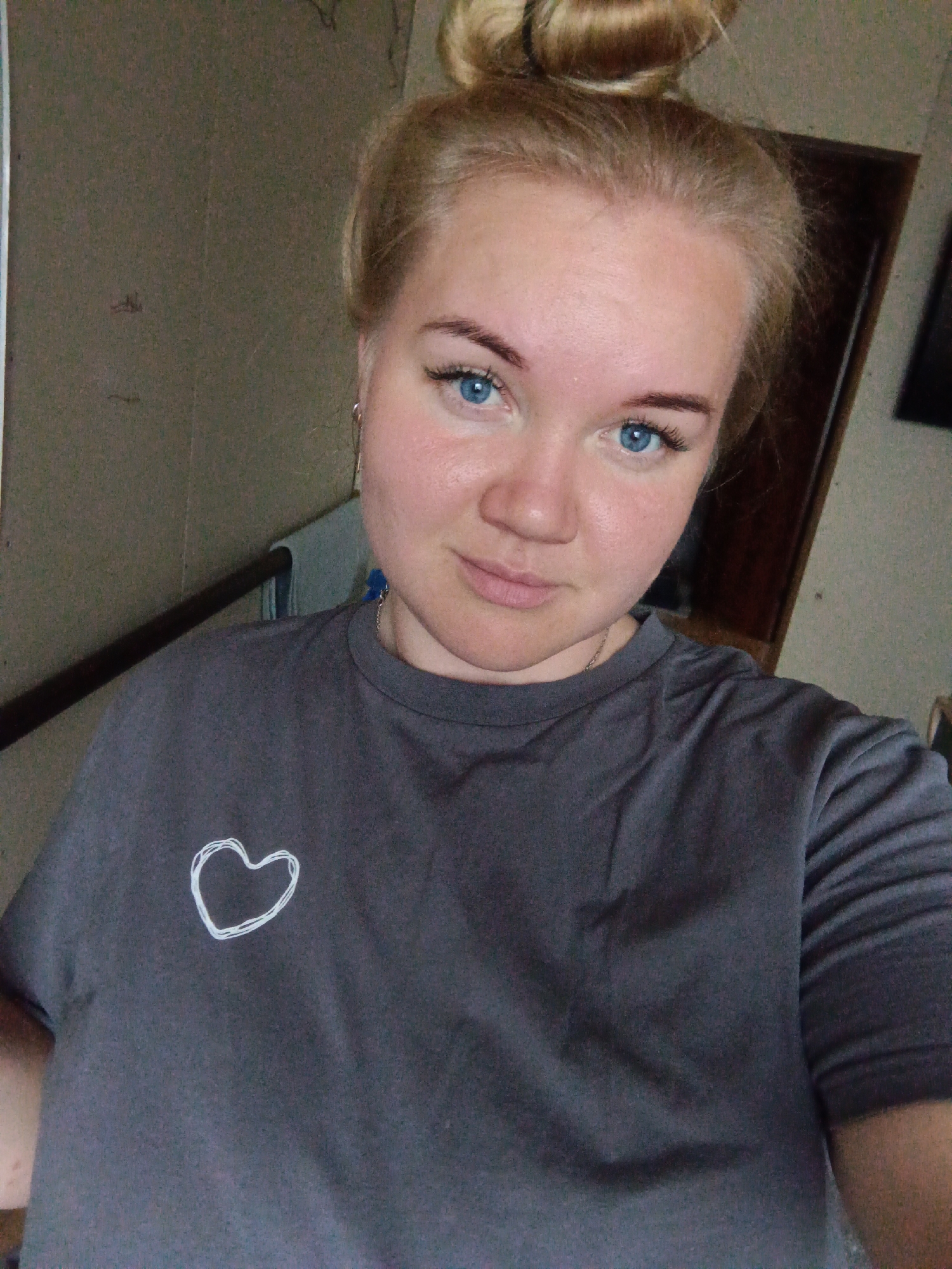 A young Caucasian woman with blonde hair pulled into a bun is taking a selfie in what appears to be a dimly lit room, possibly a hallway, given the presence of a railing and a wall texture resembling wallpaper or paint. She is wearing a dark gray t-shirt with a white outline of a heart on the left side of the chest. The woman has bright blue eyes, noticeable eyelashes, and fair skin with a slight blush on her cheeks. She is looking directly at the camera, and her expression is neutral. Behind her, a dark wooden door or furniture can be seen. The lighting in the image is somewhat harsh, creating shadows and highlights on her face. The overall environment suggests an indoor setting, potentially a residential area. There is no indication of the specific location in the city of Fokino, Russia.