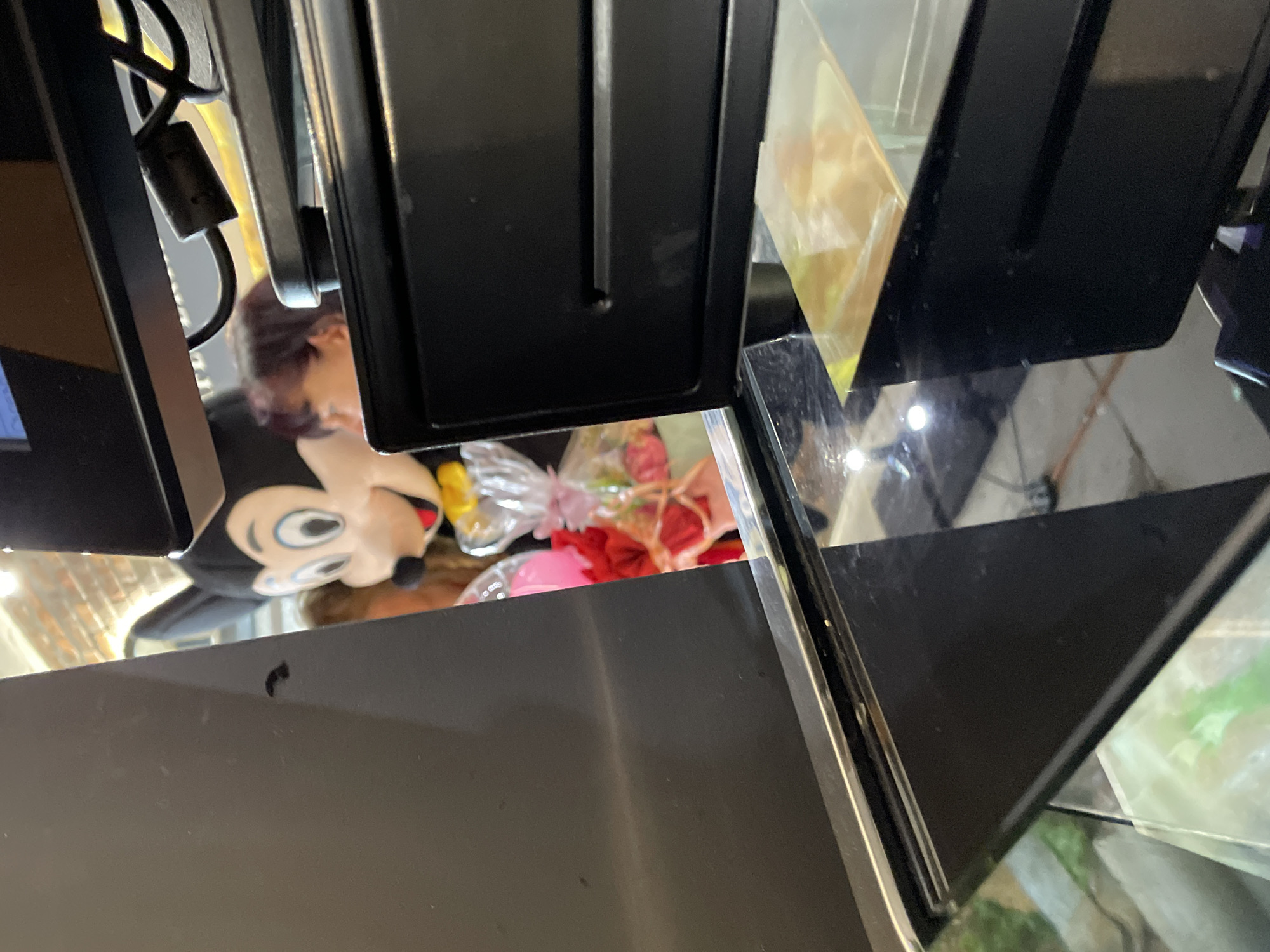 The image shows a partial reflection of a person holding a Mickey Mouse plush toy and a floral arrangement, presumably inside a store. The reflection is visible on a shiny, dark surface, possibly a display counter. The foreground features black, geometric structures, likely parts of display fixtures or shelves. The scene is likely located in a retail setting, but the image provides no further details to definitively place it in Veliko Tarnovo, Bulgaria.