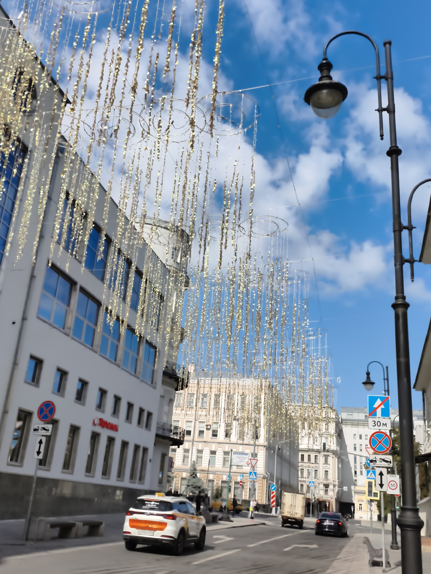 The image depicts a street scene in a city, likely in Russia based on the signage. The street is lined with multi-story buildings on the left, one of which has a white facade and many blue-tinted windows. Suspended overhead are numerous strings of golden decorative lights, creating a festive atmosphere. A black street lamp with a curved arm and a rounded light fixture is prominent on the right side of the image. Traffic signs are visible, including a red circle with a blue cross, a blue square with a white arrow, and other regulatory signs. A white taxi with orange accents is driving on the road. Several other vehicles, including a truck and a dark car, are also present. The sky is blue with scattered white clouds. There are a few people walking on the sidewalk. The overall impression is of a bright, urban environment decorated for a celebration or special occasion.