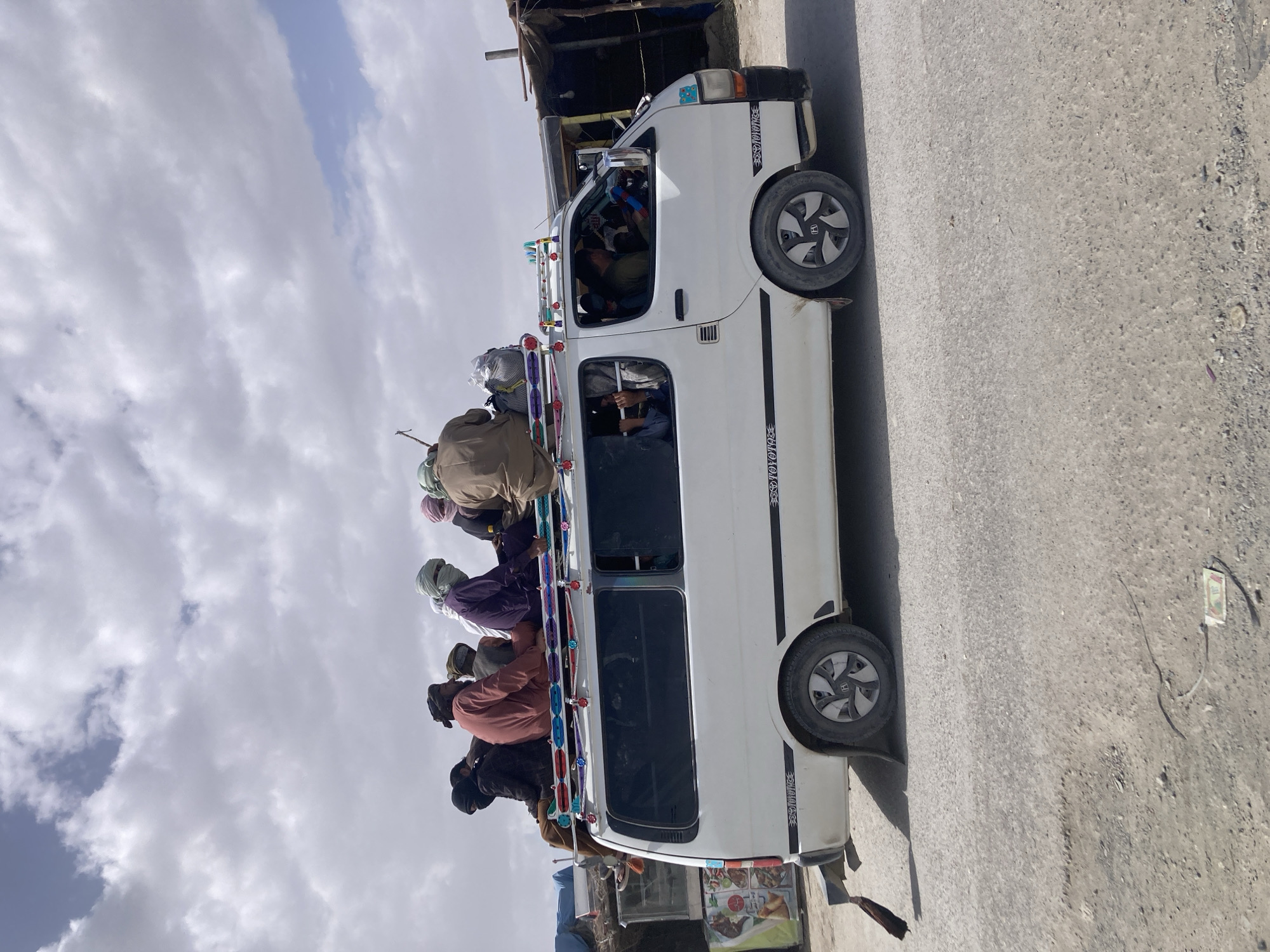 A white van travels on a paved road in Gulistan, Pakistan, heavily laden with passengers both inside and on the roof. Approximately seven individuals are positioned on the roof, while the vehicle's interior also appears fully occupied. The exterior of the van features decorative elements, including colorful lights along the roof's edge. The setting suggests a densely populated or transit area within the city.