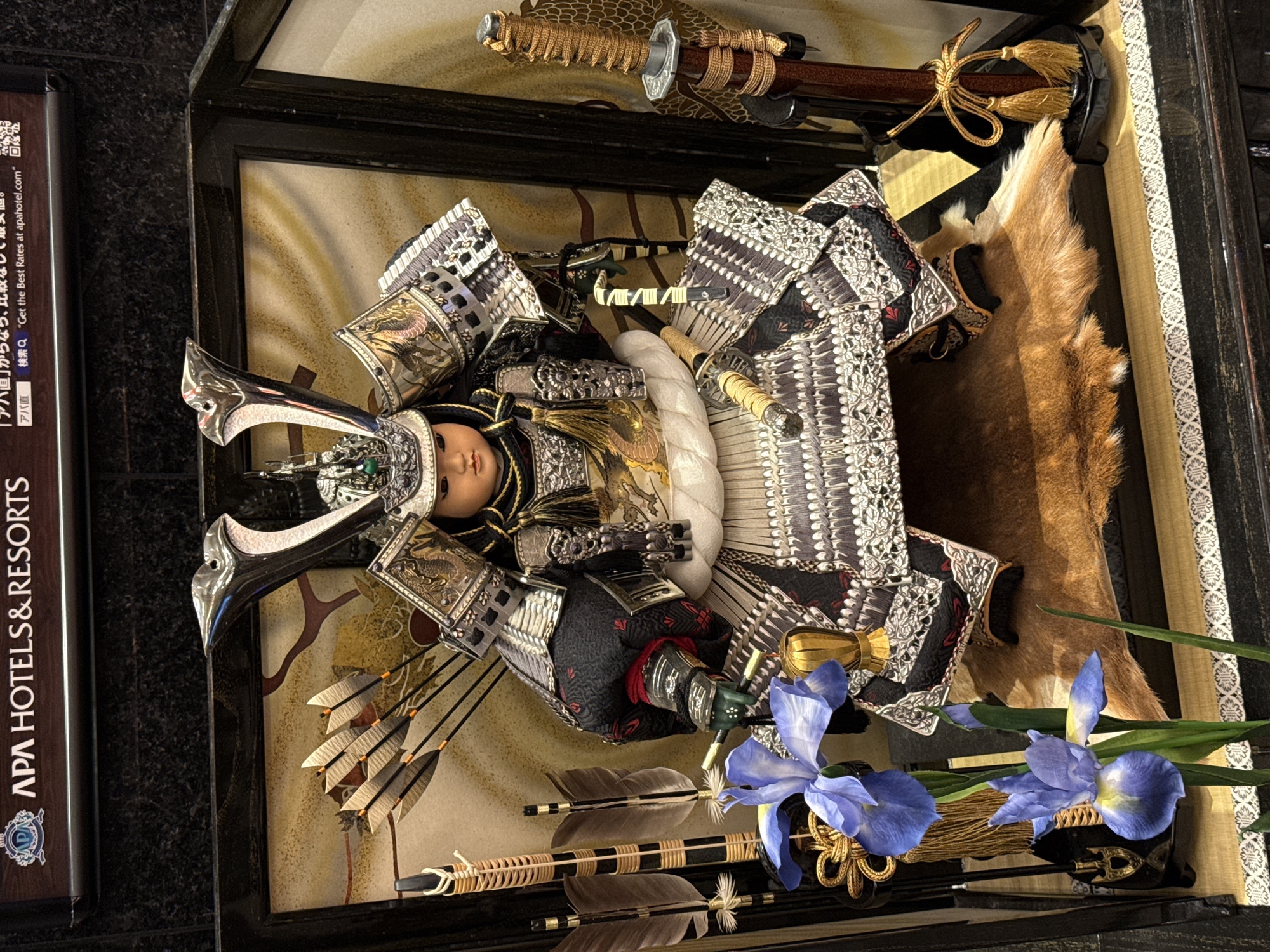 The image shows a display case featuring a traditional Japanese doll dressed in samurai armor, along with various weapons, and is located in Unknown, Unknown. The doll is the focal point, elaborately adorned with a silver helmet and intricate armor details. Beside the doll are multiple arrows, crafted with feathered fletchings, and a katana with an ornately wrapped handle. A piece of animal fur is also part of the display, adding to the traditional aesthetic. In the foreground, a spray of purple irises adds a touch of color to the scene. The background of the case consists of a textured beige and gold pattern. Additionally, the edge of an "APA HOTELS & RESORTS" advertisement is visible.