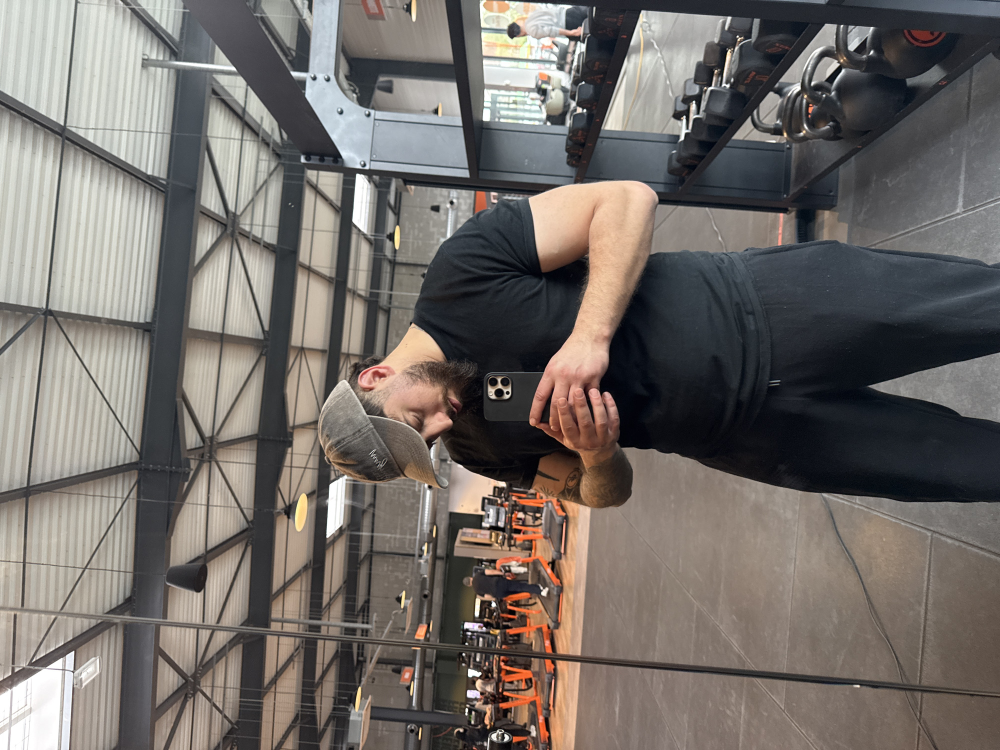 The image displays a male individual capturing a mirror selfie within a Basic-Fit gym located in Hœnheim, France. The subject, a man estimated to be in his mid-20s to 30s with a dark beard, wears a grey baseball cap, a black short-sleeved t-shirt, and black sweatpants. Tattoos are visible on his left arm. He holds a dark-colored smartphone horizontally with both hands, his gaze directed downwards at the device.
The gym environment features a dark grey tiled floor and an industrial-style ceiling composed of exposed dark metal trusses, beams, and white corrugated panels. Multiple black conical pendant lights are suspended, and rectangular skylights are visible. To the right of the subject, a black metal rack holds various black dumbbells with orange weight markings and several black kettlebells. In the reflection on the left side of the mirror, numerous orange and black treadmills are arranged, with at least one individual actively exercising on a treadmill. Further in the background, through a glass partition visible in the reflection, another person wearing a light-colored shirt is observed standing.