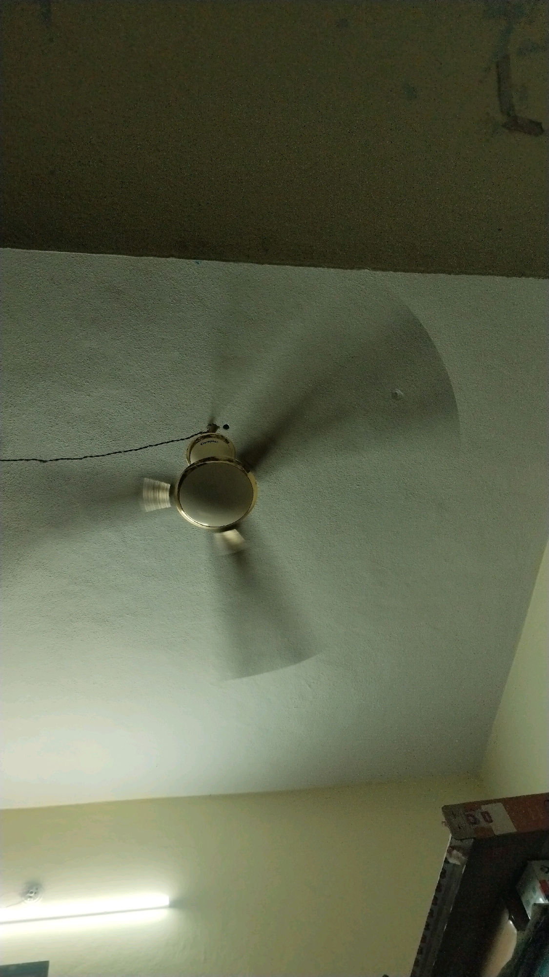 This image depicts an indoor scene, likely a room in a residential building, possibly in Hyderabad, India. A ceiling fan, appearing to be in operation due to motion blur on its blades, is centrally located in the upper portion of the frame. The fan is a light color, possibly gold or cream, with a central motor housing. It is suspended from a white ceiling by a thin wire. The ceiling itself has a textured finish and a subtle curve, casting a soft shadow from the fan. The background transitions to a darker, possibly olive-green, surface at the very top of the image. The lower part of the image shows a wall painted in a pale yellow hue, illuminated by a fluorescent tube light fixture positioned horizontally on the left. To the right, a portion of a dark wooden shelf or cabinet is visible, with some items stacked on it, appearing to be boxes. The lighting suggests a low-light environment, with the fluorescent light providing the primary illumination in the lower section. There are no people visible in the image.