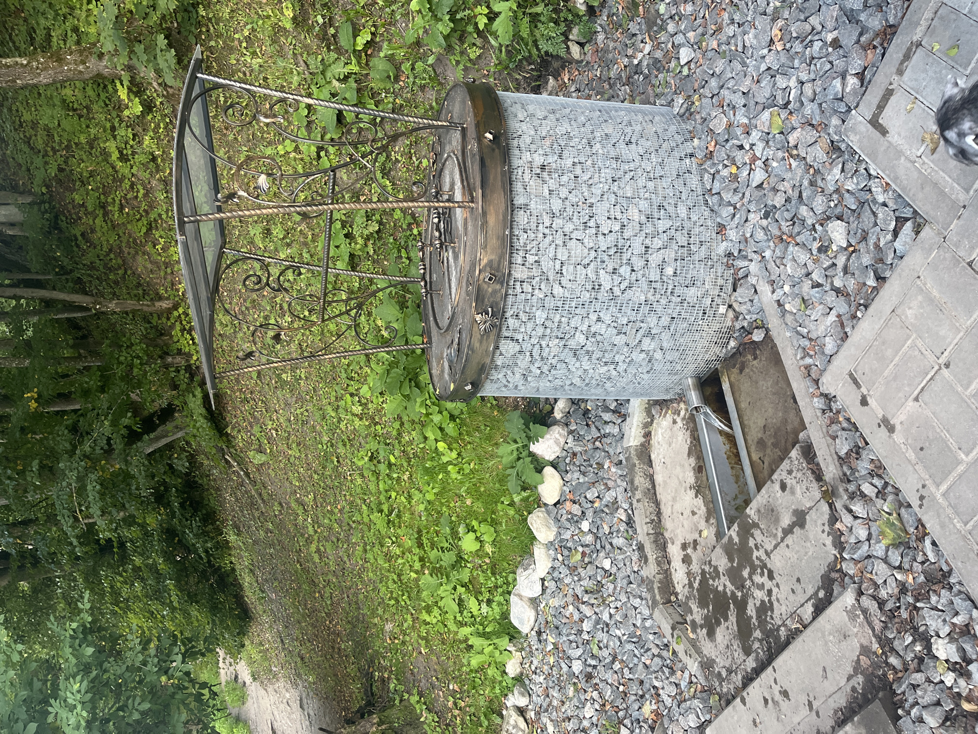 The image shows a natural spring with a stone-filled gabion structure and a decorative metal canopy in Lahojsk, Belarus. The spring is situated on a sloping, wooded area with lush green vegetation. A paved path is visible in the upper left quadrant of the image, leading up the slope. The spring itself is surrounded by gravel and stone. Water flows from the gabion structure through a metal channel into a concrete basin. There is no visible human presence in the frame.