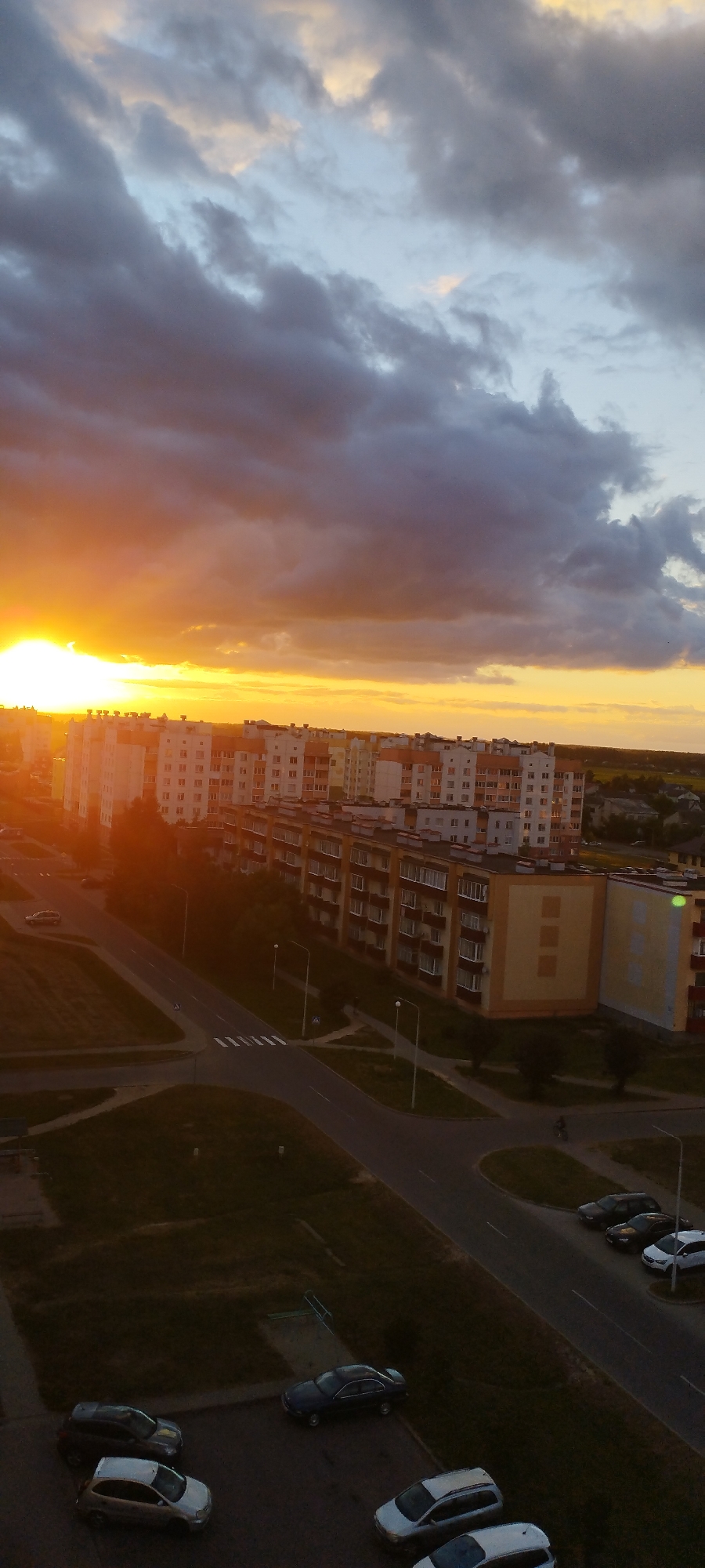 This is a vertical, high-angle photograph taken in the city of Skidzieĺ, Belarus, during sunset. The sky is filled with dramatic clouds, illuminated by the setting sun which casts a bright orange and yellow glow on the horizon, particularly on the left side of the frame. In the foreground, multi-story apartment buildings dominate the landscape, characterized by their light-colored facades and rows of windows. A paved road with pedestrian crossings winds through the scene, with several parked cars visible in a lot below. The overall impression is of a residential urban environment during a beautiful evening sky.