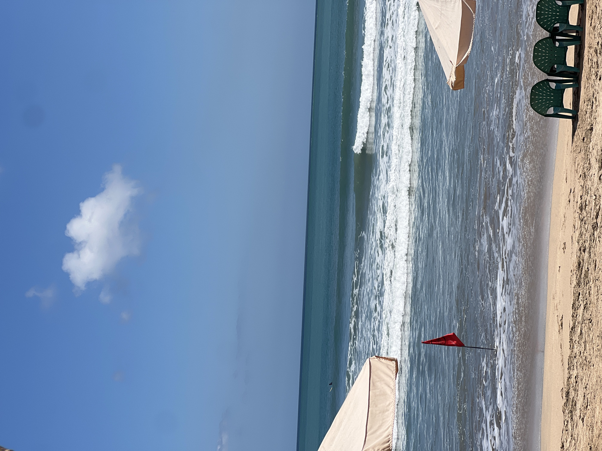 The image depicts a serene beach scene in Kuta, Indonesia, under a clear blue sky with scattered white clouds. The ocean exhibits gentle waves breaking near the shore, creating a frothy white edge. In the foreground, a sandy beach is visible, with three empty green plastic chairs positioned facing the ocean. To the left, a portion of a beige beach umbrella is framed. Closer to the water, a single red flag on a dark pole stands partially submerged in the incoming tide, indicating a warning. In the middle distance, a lone surfer can be seen on a surfboard in the water, catching a wave. The overall impression is one of a calm, inviting day at the beach.
