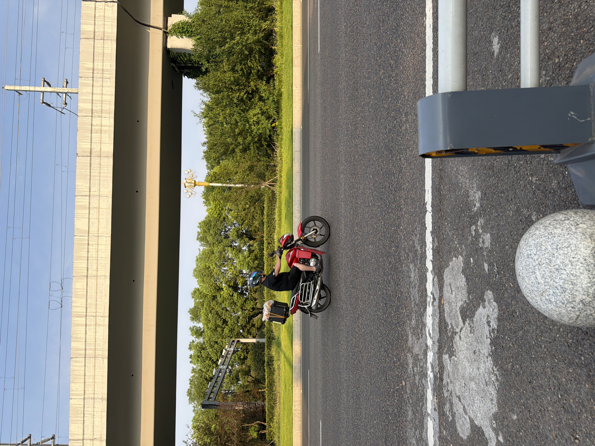 In the city of Jiangjin, China, a person wearing a black t-shirt, shorts, and a blue helmet is riding a red motorcycle on a paved road. The motorcycle has a black storage box attached to the back, containing what appears to be groceries. The rider is moving from left to right across the frame. The road is bordered by a grassy median and lush green trees. Overhead, a concrete overpass runs horizontally across the upper portion of the image, with electrical wires visible above it. The sky is clear and blue. In the foreground, a large, round, granite bollard is positioned to the left, and a grey barrier with yellow reflectors is to the right. The lighting suggests it is either early morning or late afternoon, casting long shadows.