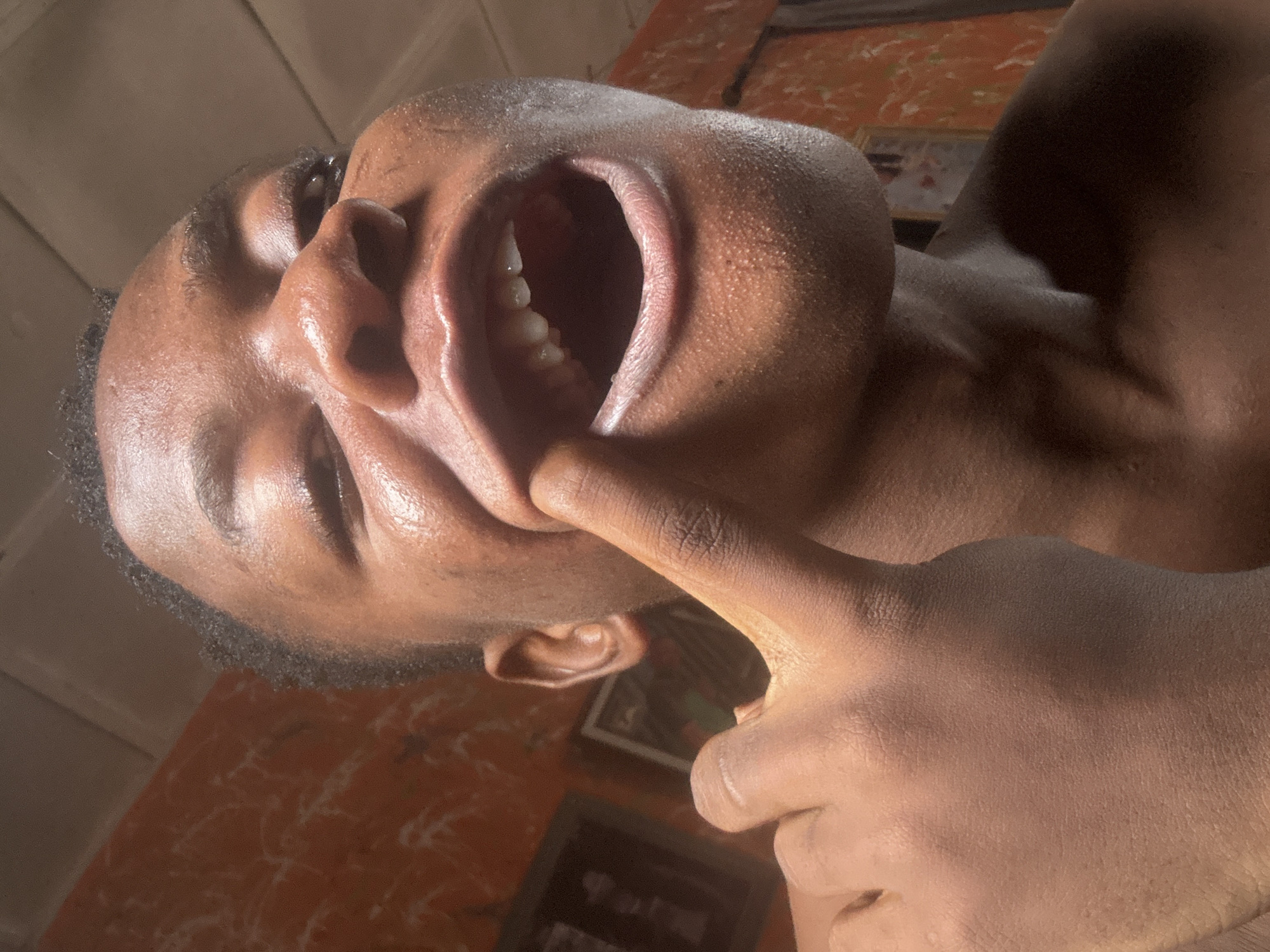 A close-up, low-angle shot captures a young Black person indoors, likely in Ikorodu, Nigeria, based on the residential setting suggested by the textured orange wall and framed artwork in the background. The individual is looking directly at the camera with their mouth open and a finger is extended, touching their teeth. Sunlight illuminates the left side of their face, highlighting their skin and the texture of their lips and the interior of their mouth, where several teeth are visible. The person appears to be in a casual pose, with their bare chest and shoulder visible. The overall lighting creates a warm and intimate atmosphere.