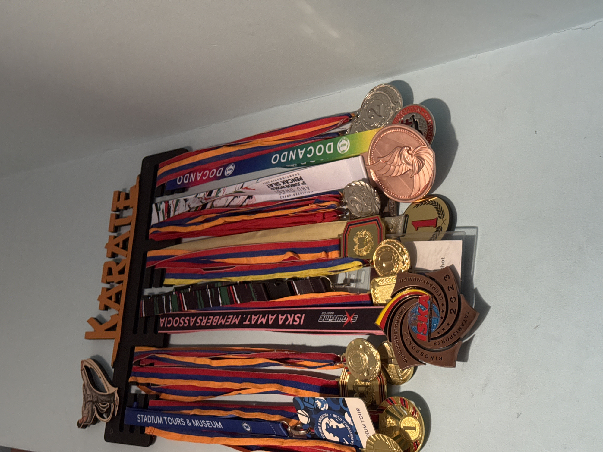 This image captures a medal display rack mounted on a light blue wall, likely in an indoor setting in Yerevan, Armenia. The dark wooden or plastic rack is shaped to vertically spell "KARATE" along its left side and features numerous hooks or slots from which a collection of achievement medals hang.
The display is densely packed with medals of various shapes and colors, including gold, silver, and bronze tones, some featuring numbers like "1" or "2" indicating placement. These medals are attached to a vibrant array of ribbons, many sporting red, blue, orange, and yellow stripes.
Visible text on the ribbons and medals provides clues to the achievements showcased. "DOCANDO" is prominently displayed on several ribbons. Another ribbon mentions "5 JUNIOR WORLD PENCAK SILAT CHAMPIONSHIP 2014 ABU DHABI," indicating an international martial arts competition. Other text includes "ISKA AMAT. MEMBERS ASSOCIA," "SHOWTIME SPORTS," and "TATAMISPORTS" on ribbons. A distinctive star-shaped medal clearly reads "RINGSPORTS," "TATAMISPORTS," "WORLD CHAMPIONSHIPS," "GERMANY/MUNICH," and "2023," suggesting a recent international event. Further down, ribbons and a lanyard display "STADIUM TOURS & MUSEUM" and "SA FOOTBALL CLUB DIUM TOUR," indicating participation or visits to sports venues. The collection suggests a background of extensive involvement in sports, particularly martial arts like Karate and Pencak Silat, with achievements spanning several years and international locations.