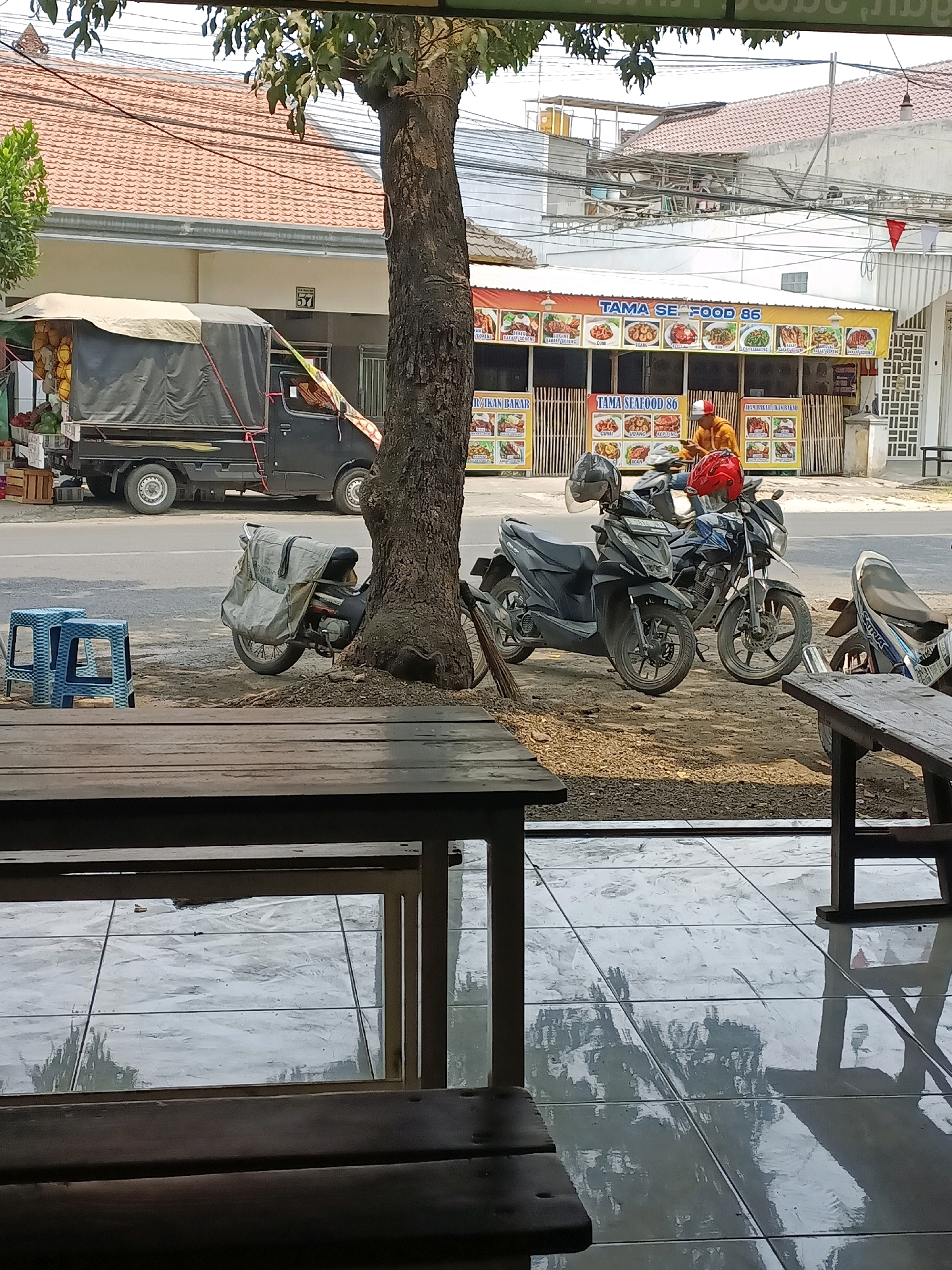 The image depicts a street scene in Sukomulyo, Indonesia, likely in front of a food stall. In the foreground, wooden tables and chairs are visible, suggesting an outdoor seating area for a restaurant. Beyond the seating area, a gray pickup truck is parked on the side of a road, its cargo bed covered with a dark tarp. The truck appears to be selling produce, with fruits like durians and papayas visible at the back.
Further down the road, a food stall named "TAMA SEAFOOD 86" is prominent, with colorful banners displaying various seafood dishes. A person wearing an orange jacket and a red and white cap is seated on a motorcycle in front of the stall, seemingly engaged with a mobile phone. Several motorcycles are parked in front of the stall. The environment shows typical street elements with buildings, power lines, and trees lining the road. The ground appears to be paved with tiles in the foreground and dirt/gravel in front of the food stall.
