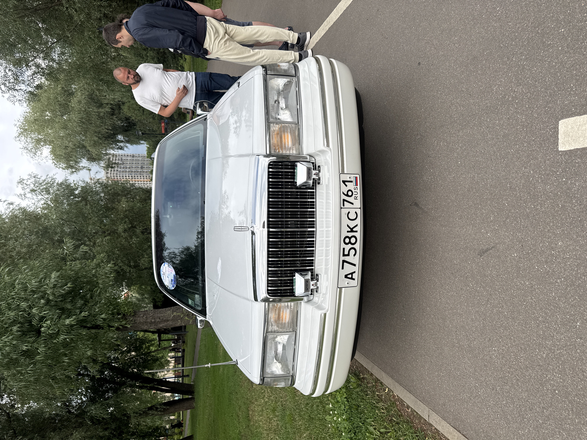 Two men are standing next to the front of a white Lincoln Town Car parked on an asphalt path next to a grassy area with trees. One man, wearing a white t-shirt and blue jeans, is standing and looking towards the car. The other man, wearing a dark jacket and light-colored pants, is standing behind the first man, also looking down at the car. The scene appears to be outdoors in a park or public space, with trees and some buildings visible in the background. The time of day seems to be daytime, and the weather appears overcast. A license plate on the car reads "A758KC 761 RUS". The car has a prominent chrome grille and headlights.