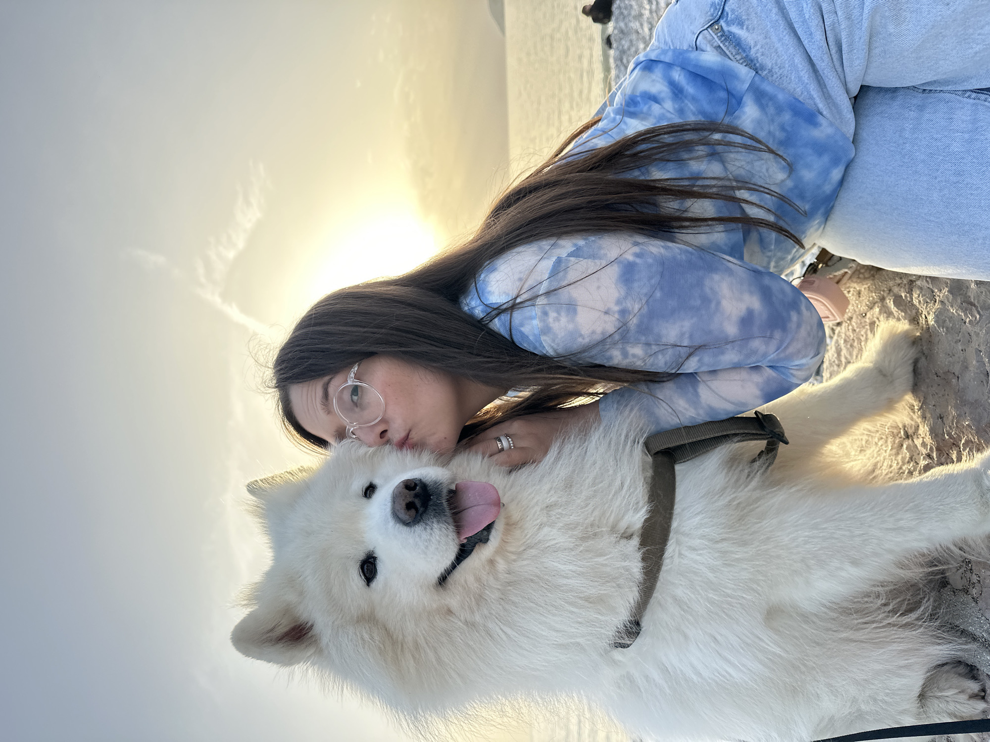 A young woman with long brown hair and glasses is kissing a large white dog. The dog is wearing a harness and has its tongue sticking out. They are outdoors, possibly at a beach or rocky coastline, at sunset in Šušanj, Montenegro. The sun is setting in the background, and the sky is a mix of yellow and grey. The woman is wearing a blue and white tie-dye sweatshirt and jeans.