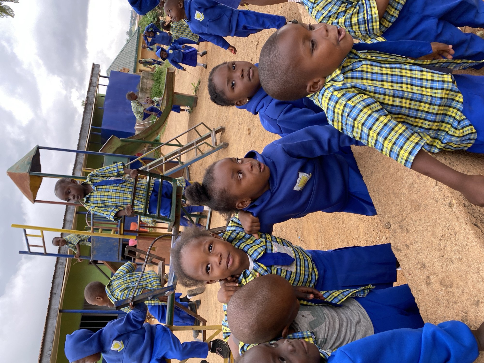 A group of children in blue uniforms are playing on a playground in Gura Top, Nigeria. Some children are on the playground equipment, including a slide and swings. Other children are standing or sitting around the playground. The environment appears to be a schoolyard. Buildings and vegetation are visible in the background.