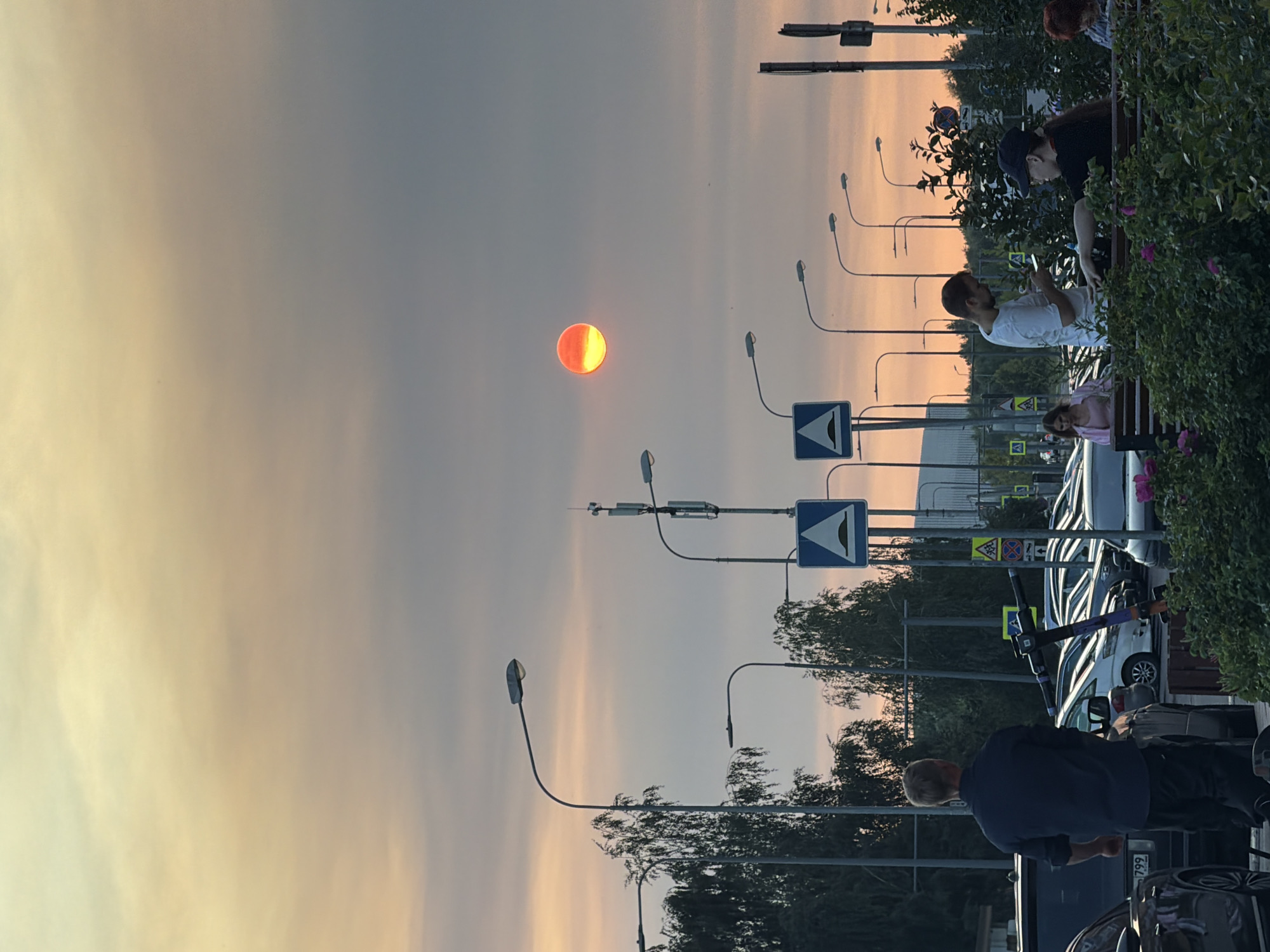 The image depicts a street scene in Shcherbinka, Russia, during sunset. The sky is filled with hazy, orange and yellow clouds, obscuring the sun which appears as a bright, reddish-orange orb with a lighter yellow section. Several streetlights line the road, with one on the far right appearing to have a communication antenna attached. Road signs are visible, including a blue sign with a white triangle indicating a speed bump and another yellow diamond-shaped sign with a pedestrian crossing symbol.
In the foreground, a man in a dark blue shirt and dark pants stands facing away from the camera, looking towards a row of parked cars. To his left is a dark colored SUV with a license plate ending in "799". To his right is a parked electric scooter.
Further into the scene, a group of people are gathered around a wooden bench surrounded by lush green bushes with pink flowers. A man in a white t-shirt is looking at his phone while standing next to a woman wearing a black t-shirt and a baseball cap. Another woman with red hair is seated to the right. Several cars are parked in the background, and trees with thin trunks and dense foliage are scattered throughout the scene. The overall impression is a public urban space at dusk, with elements of both infrastructure and human activity.