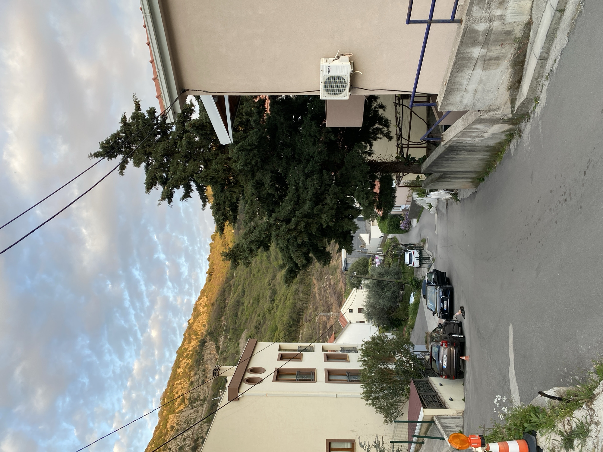 The image captures a street in Budva, Montenegro, showcasing a residential area nestled against a hillside. The street is lined with buildings of varying heights and architectural styles. In the foreground, a building with multiple windows and a brown trim is visible. Adjacent to it, a tree partially obscures the view of the road. Further up the street, cars are parked, and a person is standing near one of the vehicles. The road extends into the distance, revealing more buildings and vegetation. In the background, a hill covered in greenery rises towards the sky, which is filled with clouds. Utility lines run across the top portion of the frame. A building with an air conditioning unit is positioned on the right side of the street. An orange and white traffic cone is visible near the bottom of the image.