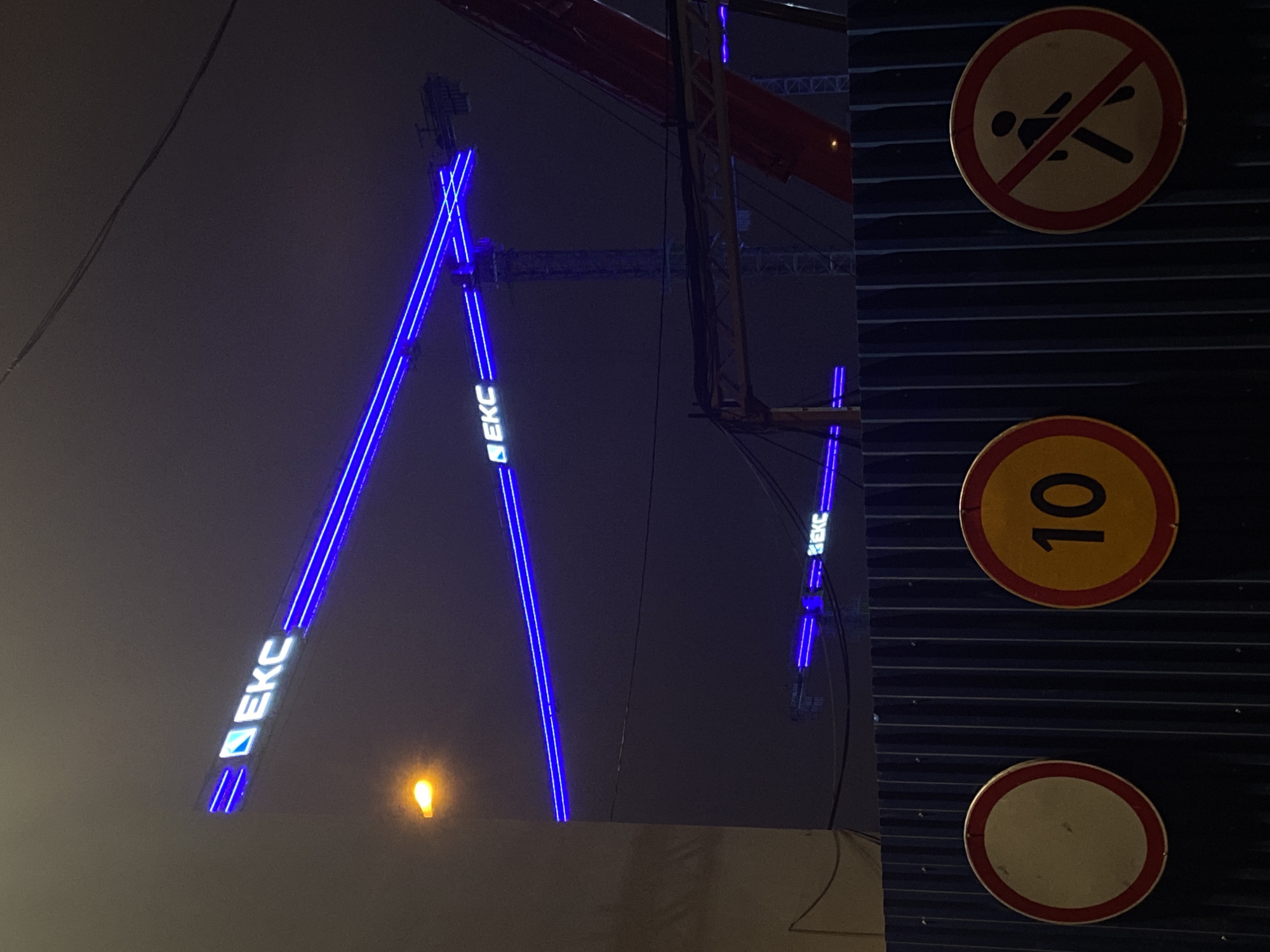 The image shows a nighttime scene in Moscow, Russia, featuring construction elements and road signs. A large construction crane with the illuminated logo "EKC" is prominent in the background. A set of three road signs is visible on a vertical structure on the right. The top sign depicts a crossed-out pedestrian, the middle sign displays the number "10", and the bottom sign is blank. A streetlight illuminates the crane from below. The sky is overcast.