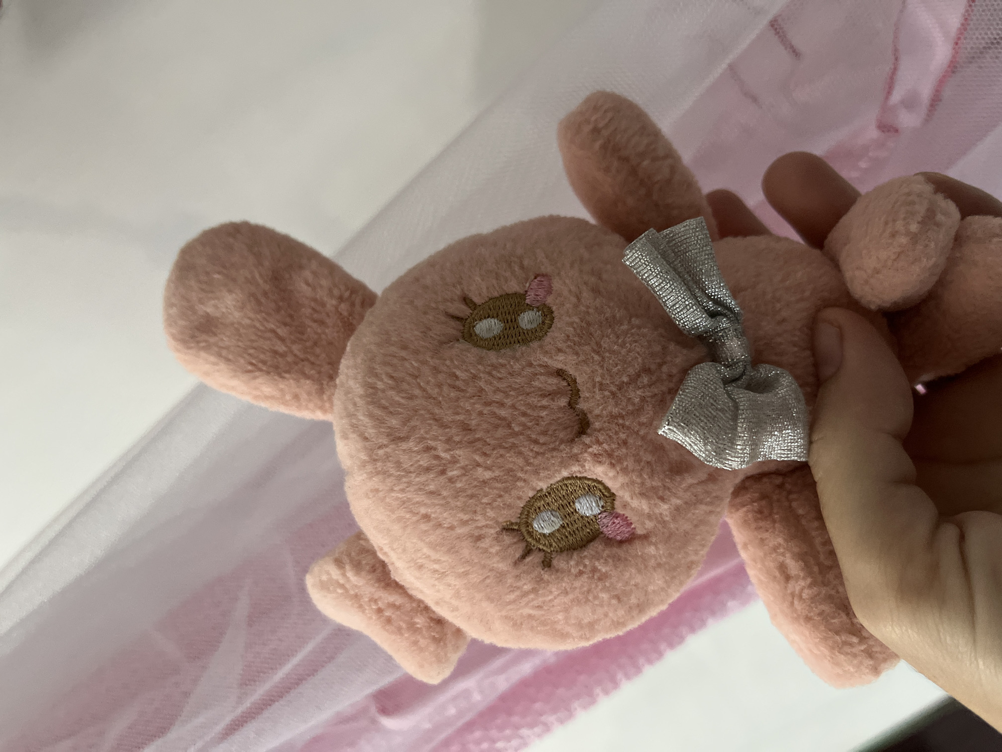 The image captures a close-up of a small, pink, fluffy plush toy being held by a human hand. The toy features embroidered facial details, including two closed eyes with brown outlines, white highlights, and small eyelashes, along with a tiny pink tongue playfully sticking out. A shiny, silver-colored bow is tied to the top of the toy's head.
A light-skinned hand is visible on the right, gently gripping the plush toy. The background consists of soft, sheer fabric in shades of light pink and white, resembling tulle or netting, with hints of a solid pink fabric layer underneath.
The primary activity is a hand holding the plush toy. The toy's embroidered expression suggests happiness or contentment. No text is visible in the image. The setting appears to be indoors. The location context for this media is Lhokseumawe, Indonesia.