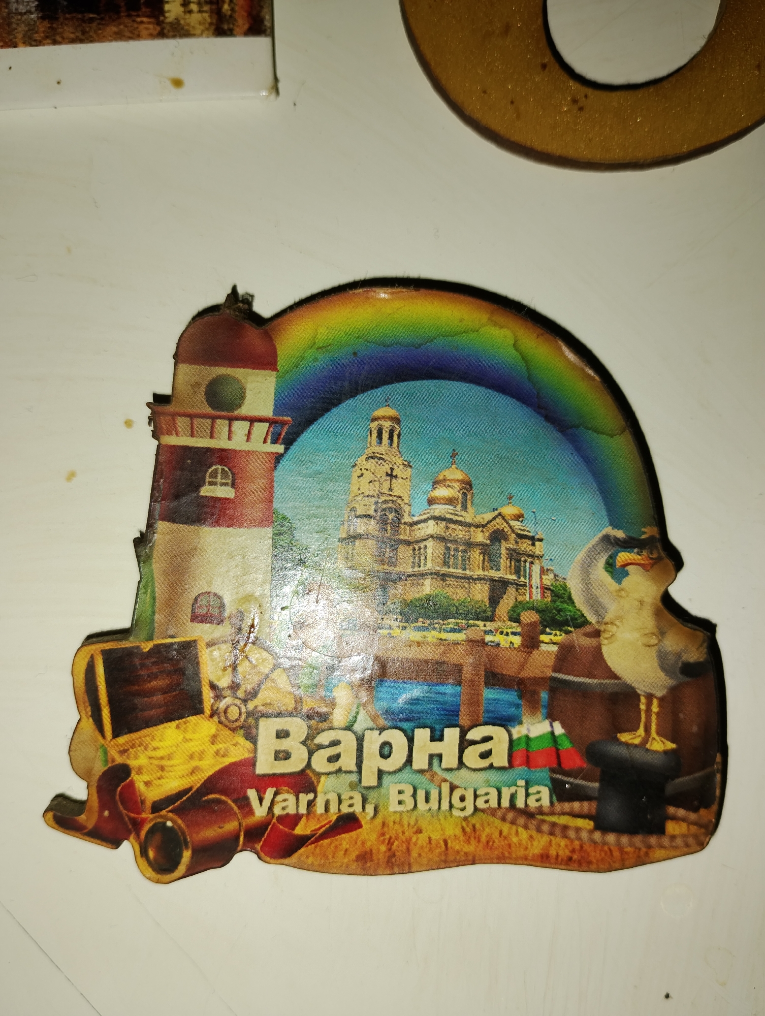 A souvenir magnet depicts a scene with a lighthouse, a cathedral, a seagull, and a treasure chest, along with the text "Варна, Varna, Bulgaria." The lighthouse is red and white, and the cathedral is gold-domed. The seagull is depicted wearing a hat and saluting. The treasure chest is open and appears to be filled with gold coins. The background shows a rainbow and a body of water, possibly a harbor. The overall image is colorful and appears to be printed on a flat, glossy surface. The magnet is mounted on a white surface.
