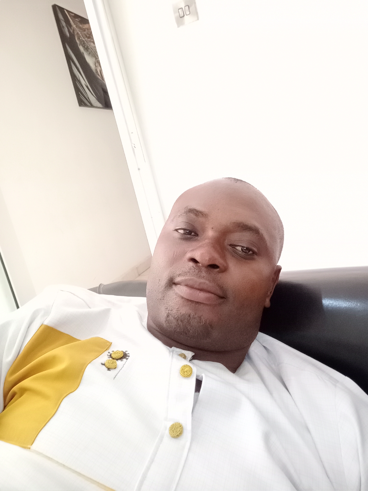 The media file presents a close-up portrait of a dark-skinned man, likely in his middle age, with a bald head and a faint stubble beard. He is seated on a dark, possibly leather, upholstered sofa or chair, looking directly at the camera with a calm and neutral expression.
The man is wearing a traditional-style garment, predominantly white, featuring a subtle textured pattern. A prominent golden-yellow panel is sewn diagonally across the left side of his chest (viewer's left). The garment has a placket adorned with several circular, golden-colored buttons, each intricately patterned. Small embroidered details, resembling stylized circular motifs, are visible near the buttons on his left side.
The background is simple, consisting mainly of a light-colored wall, likely white or off-white. To the left, a dark-toned framed picture or artwork is mounted on the wall. To the right, a white vertical structure, possibly a door frame or wall edge, is visible. In the upper right corner, a light-colored object with two distinct "00" markings, possibly an electrical fixture or part of an alarm system, is present. The lighting is bright and even, indicative of an indoor setting during the daytime. This scene is located in Gwarinpa, Nigeria.