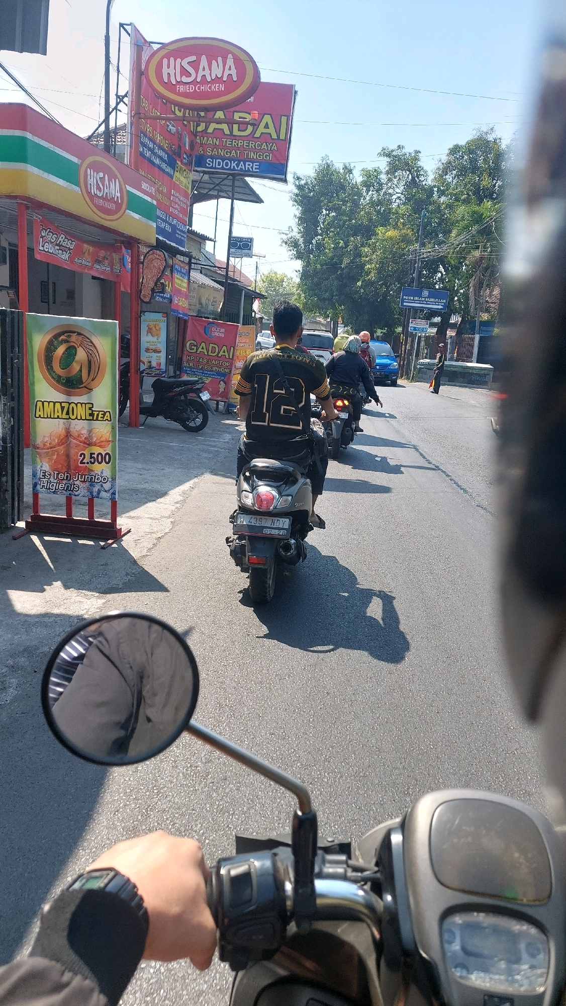 This is a first-person view from a motorcycle traveling on a street in Sidoarjo, Indonesia. The motorcycle is in motion on an asphalt road under a clear blue sky. In the foreground, the rider's gloved left hand is visible on the handlebar, and the motorcycle's rearview mirror reflects the rider's arm and shoulder.
To the left of the road, there are several businesses. A prominent sign for "HISANA FRIED CHICKEN" is visible, along with another smaller "HISANA" sign. A large, vertical advertising stand for "AMAZONE TEA" displays a price of "2,500" and advertises "Es Teh Jumbo Hygienis." Other signs for businesses like "GADAI" are also present.
Several motorcycles are on the road. The motorcycle directly in front of the viewer has a rider wearing a black shirt with the number "12" on the back. Further ahead, other motorcycles are visible, including a blue car in the distance. Trees line the right side of the road.
The scene appears to be a typical urban street in Sidoarjo, with commercial establishments and traffic. The time of day is likely midday, given the bright sunlight and distinct shadows.