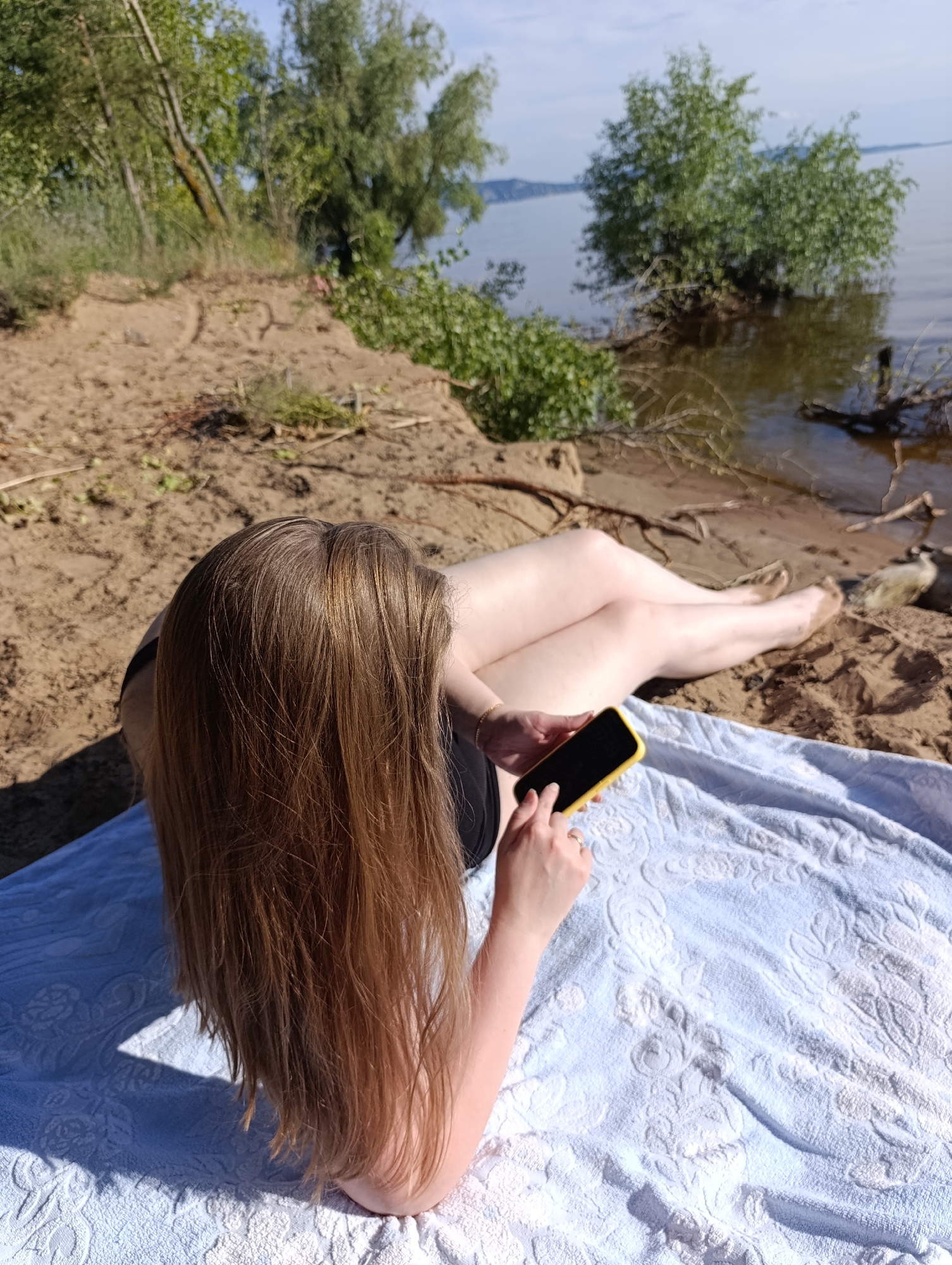 In an outdoor scene, a person is lying on a sandy bank near the water. The individual has long, blonde-brown hair and is wearing a black top. They are holding a yellow-cased smartphone in their right hand, with their left hand positioned as if interacting with the screen. The person is lying on a textured, light blue towel. The background shows a body of water, with trees on the opposite bank and some vegetation on the immediate bank. The lighting suggests it is daytime, with bright sunlight creating shadows. The overall mood appears relaxed and peaceful.