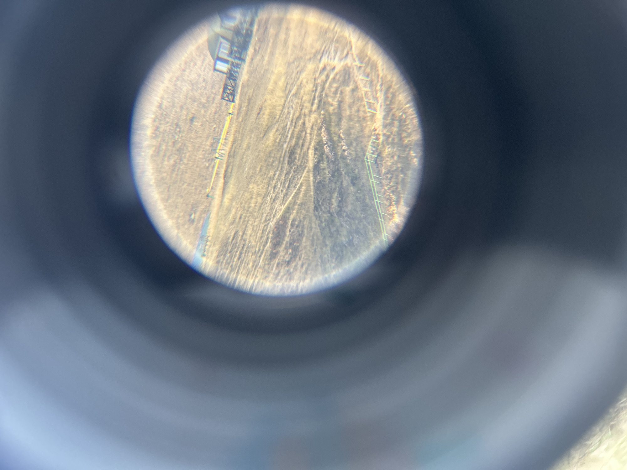 The image, viewed through a circular lens or aperture, shows a dry, grassy field with faint tire tracks. In the background, a wooden structure, possibly a building or a fence, is visible. A yellow object, possibly a ribbon or a line, runs horizontally across the field. The scene appears to be outdoors in a rural or semi-rural setting. The lighting suggests it is daytime, with the sun casting a warm glow on the landscape. The location is identified as Sakhyurta, Russia.