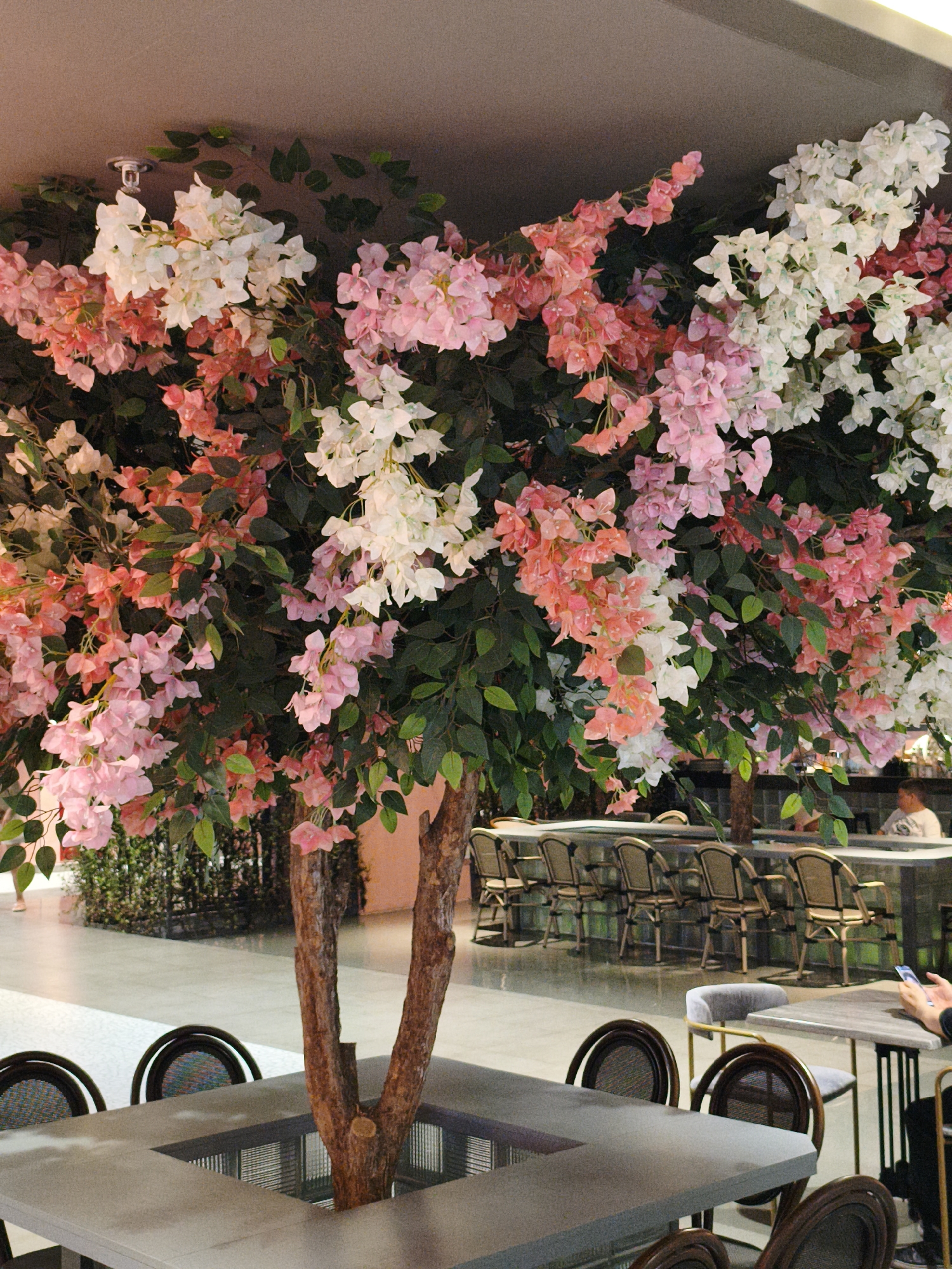 A wide-angle, eye-level shot shows a large artificial tree with pink and white flowers and green leaves in the foreground. The tree has a thick, textured trunk that splits into two main branches. The tree is centered within a modern restaurant or cafe interior. In front of the tree is a square table with a recessed section where the tree trunk sits. Several chairs with woven backs and dark frames are visible around the table. In the background, a bar area is visible with numerous stools and a counter. A few people are present in the background, including one individual sitting at the bar and another partially visible in the far right. The flooring appears to be polished concrete. The overall impression is that of an indoor dining space with a prominent decorative tree.