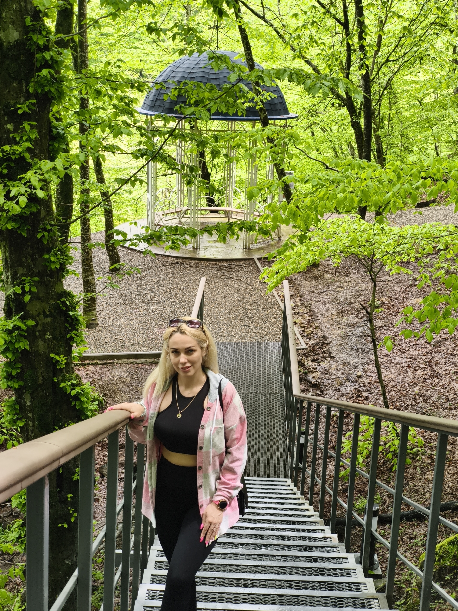 A woman is standing on metal stairs in a park in Goryachiy Klyuch, Russia. The stairs lead toward a white gazebo with a black roof. The woman is blonde and wears sunglasses on top of her head, a black crop top, black leggings, and a pink and white flannel shirt. She is holding the railing of the stairs. Trees surround the staircase and gazebo, with green leaves visible. The ground on either side of the stairs is covered in brown leaves.