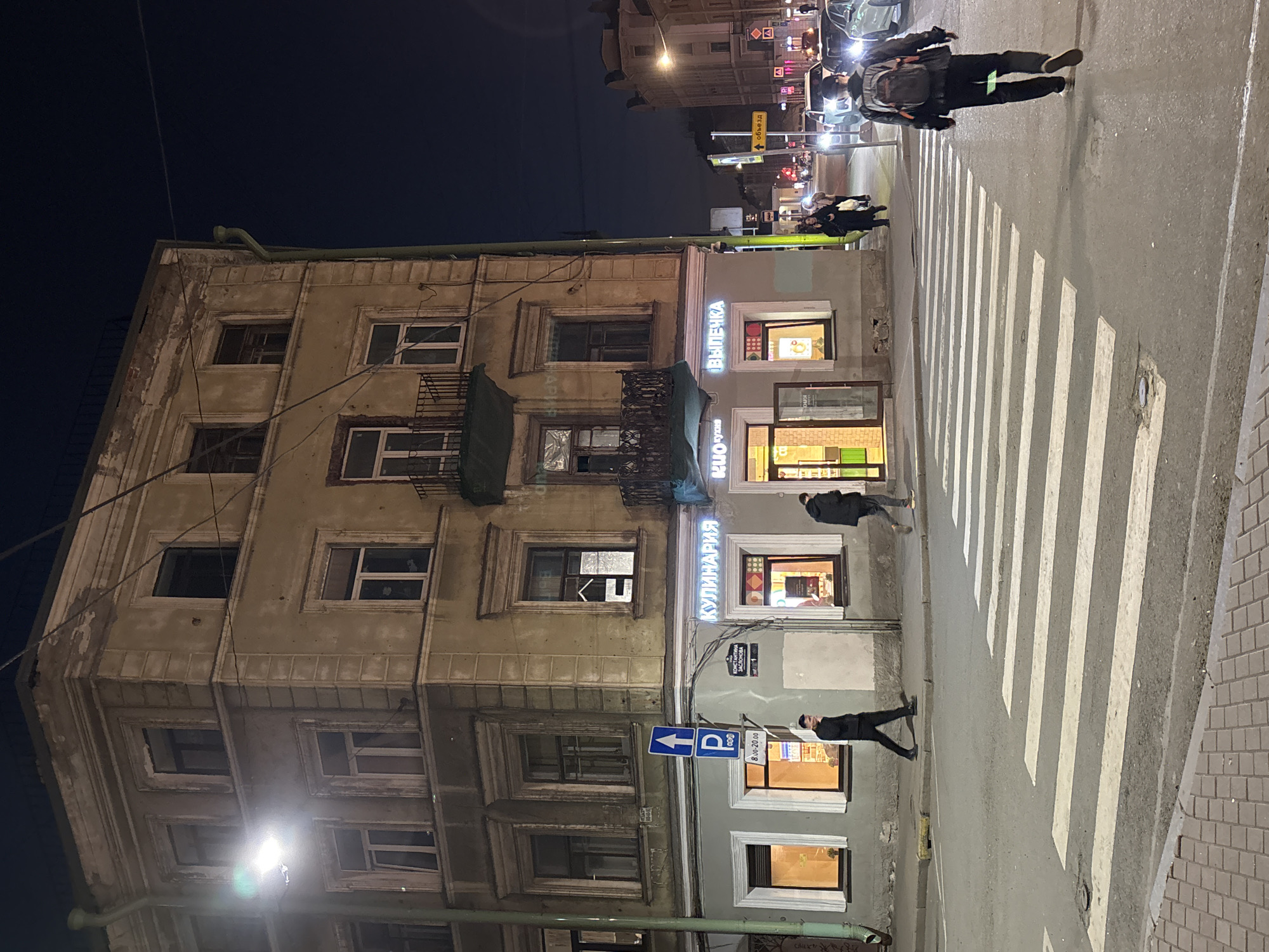 The image shows a night scene in Saint Petersburg, Russia. The left side of the frame is dominated by an old, multi-story building with faded paint, many windows, and some with balconies. The building's architecture appears dated. Adjacent to this building is a newer, well-maintained building with illuminated storefronts. Signage in Russian is visible above the windows, one of which reads "КУЛИНАРИЯ" (culinary) in neon lights and another reads "Выпечка" (pastries) in neon. A parking sign with a "P" symbol and an arrow pointing upwards is mounted on the building's facade. A pedestrian crosswalk is in the foreground, with several people crossing the street. Some traffic is visible in the distance under streetlights. The overall environment is urban, and the lighting suggests it is after dark.