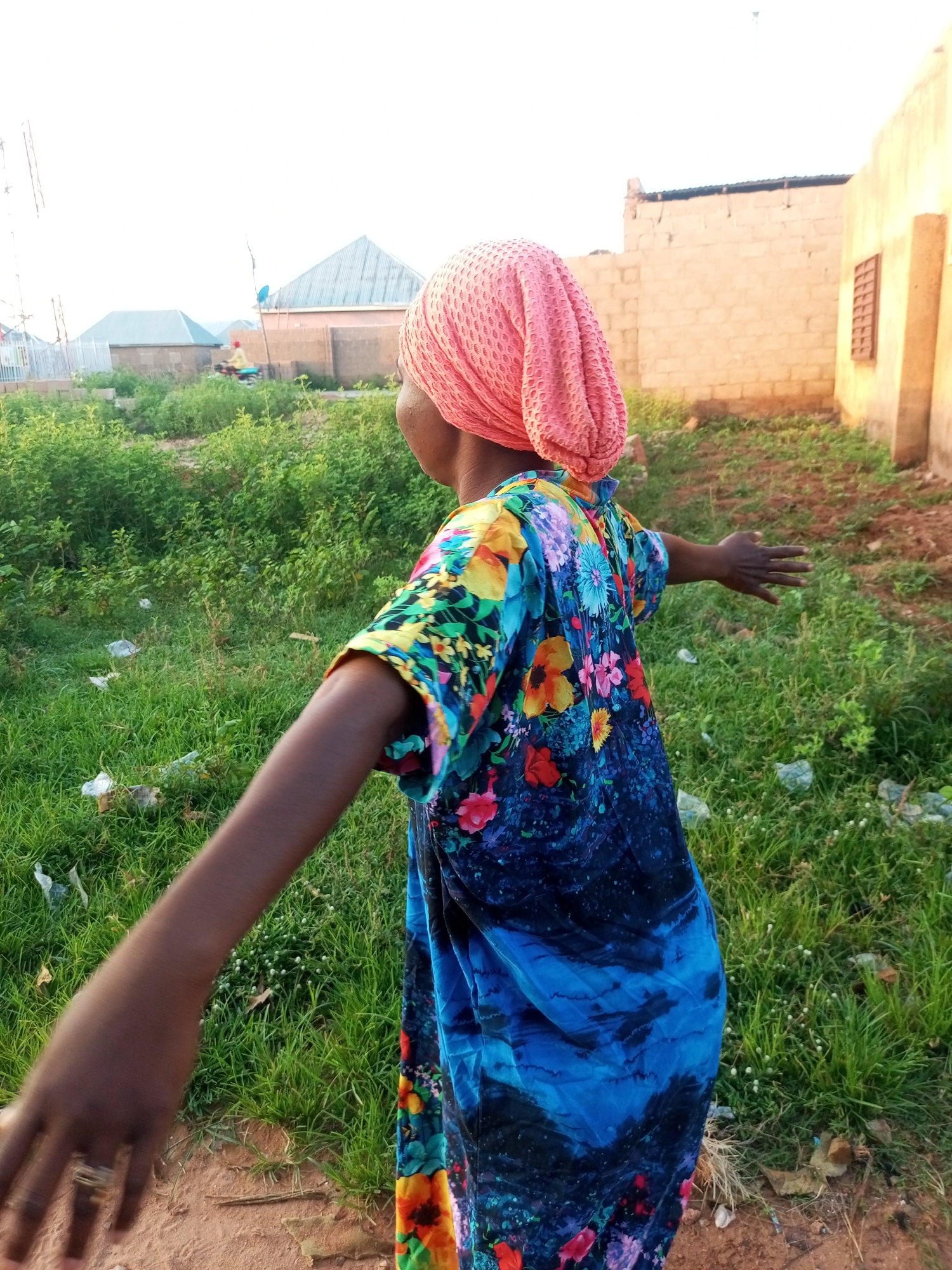 In Tempure, Nigeria, a young girl with dark skin, wearing a pink headscarf and a vibrantly colored dress adorned with floral patterns, stands in an outdoor, unpaved area. The dress features a blend of blue, black, and bright floral designs. Her left arm is extended outwards, with her hand open and facing forward. She has rings on two fingers of her left hand. Behind her, a two-story building constructed with unpainted brick is visible, with a small window on its right side. In the background, more residential structures with corrugated metal roofs can be seen, along with lush green vegetation and scattered debris. The sky is bright and overcast.