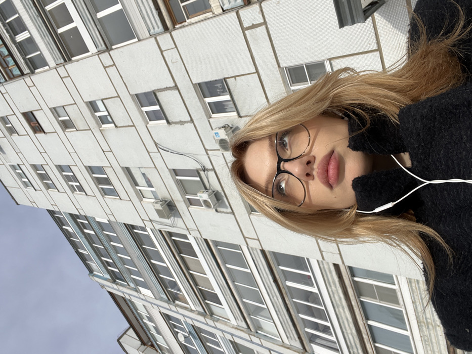 A young adult female is depicted from a low-angle perspective, looking upwards, positioned against a multi-story residential building in Chaikovsky, Russia. The subject has light-colored hair, wears black-rimmed glasses, and is dressed in a dark, textured garment, possibly a sweater or jacket. White wired earphones are visible around her neck, with the cable splitting in front of her chest. Her head is tilted slightly back, and her eyes are directed upwards and to the left of the frame. The background features a tall, light-grey residential building constructed with modular panels or small tiles, characterized by numerous rectangular windows, some with white frames, and multiple external air conditioning units affixed to its facade. Utility wires are visible descending the building's exterior. The sky above is clear and light blue, indicating a daytime scene.
