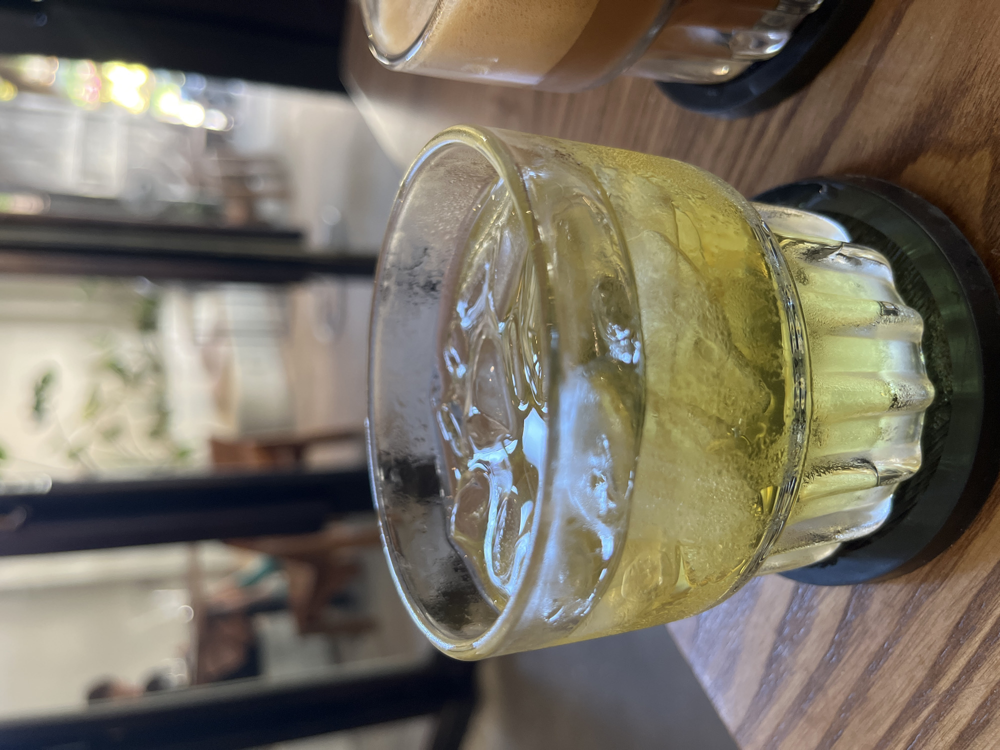 A close-up shot depicts a glass of ice water, possibly a cocktail, sitting on a black coaster on a wooden table. The ice cubes within the glass are clearly visible. In the blurred background, visible through a window or open space, are hints of an interior or outdoor setting, possibly a cafe. The location might be in Bac Nha Trang Ward, Vietnam, based on context clues. There is another glass with a dark amber liquid in the upper part of the frame, likely a coffee or other beverage. The image is taken at an oblique angle.