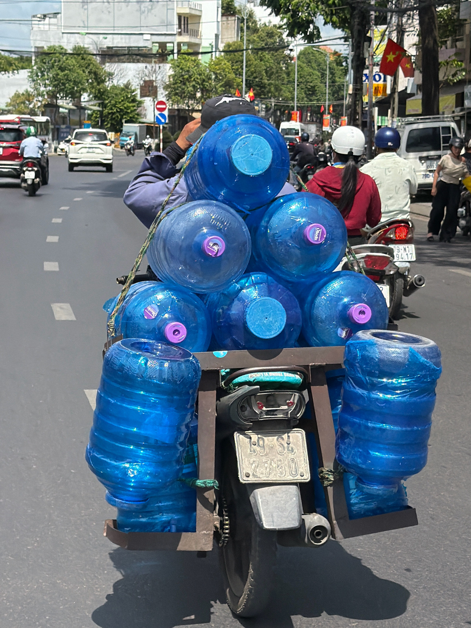 The image captures a street scene in Nha Trang, Vietnam, focusing on a person riding a motorcycle laden with multiple large blue water bottles. The person, wearing a dark baseball cap and a blue garment, is partially visible from behind, steering the motorcycle. The water bottles are stacked and secured on the rear of the motorcycle. One bottle is prominently in the foreground, displaying a purple cap and a blue and purple label. The motorcycle's license plate is visible, reading "E9-SE 2760". In the background, other vehicles, including cars and motorcycles, are present on the road, along with buildings and trees. The scene appears to be on a sunny day, with shadows cast on the asphalt road.