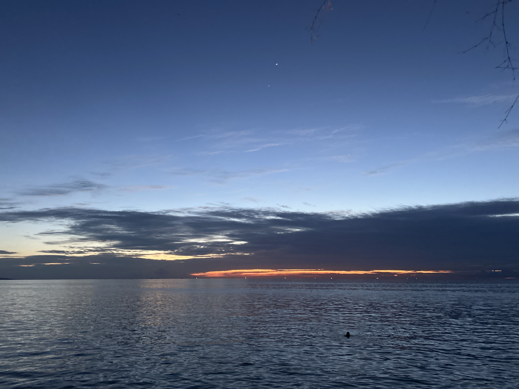 The image depicts a serene seascape at dusk over the waters of Koh Phangan, Thailand. The sky is a gradient of deep blue at the top transitioning to lighter hues near the horizon, with wispy clouds scattered across the expanse. Two bright points of light, possibly stars or planets, are visible in the upper portion of the sky. Along the horizon, a thin band of orange and yellow light from the setting sun is visible, partially obscured by dark, heavy clouds. The calm water reflects the colors of the sky, creating a tranquil atmosphere. Small lights, likely buoys or distant boats, are dotted across the water near the horizon. In the foreground, a single dark silhouette, possibly a person swimming or a small object, is present in the water. The edge of a tree branch, silhouetted against the sky, is visible in the upper right corner.