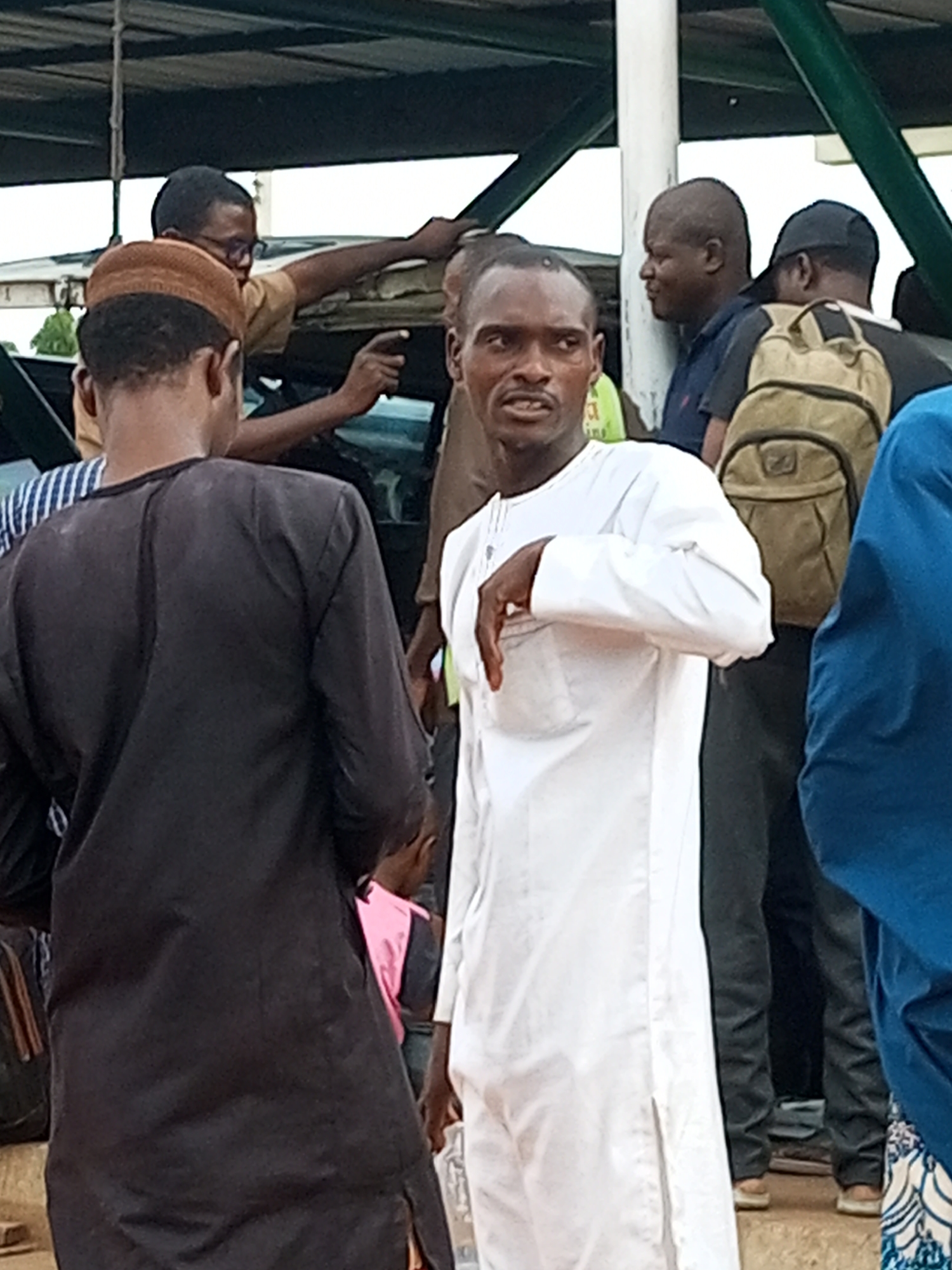 A group of people, likely male, are gathered under a covered structure in Gombe, Nigeria. A man in the foreground, wearing a white traditional garment, is looking directly at the camera with a tense expression and has his left hand raised, palm facing outward. To his left, another person in a black traditional garment, wearing a brown cap, is facing away from the camera. Behind them, several other individuals are visible, including one with a khaki backpack and another in a blue garment. The environment appears to be outdoors, possibly near a transportation hub, given the presence of a vehicle and what looks like a waiting area. The overall impression is one of a moment of interaction or confrontation.