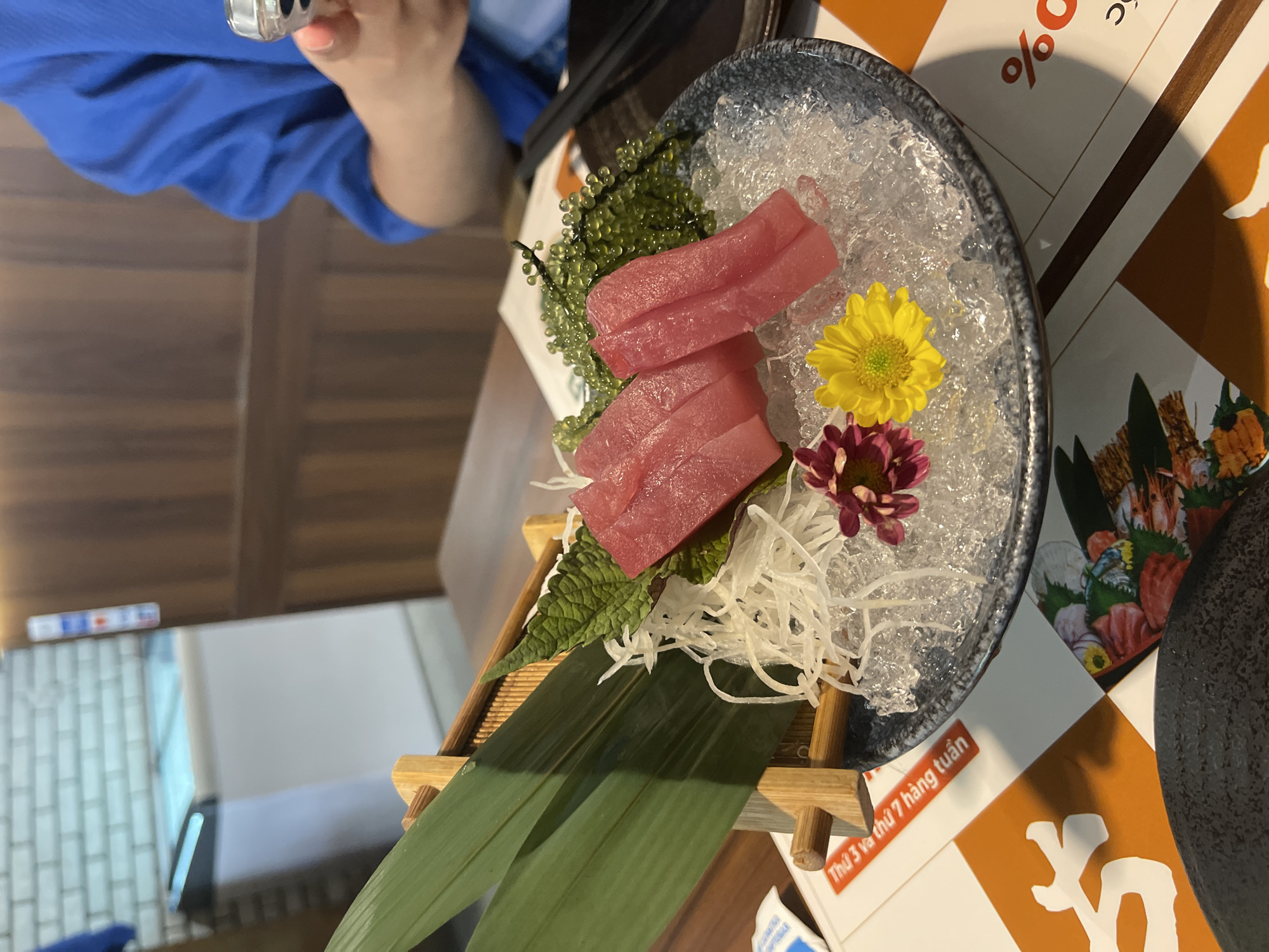 A slightly angled close-up shows a plate of sashimi atop a small, light-wood stand. The sashimi consists of sliced raw fish, possibly tuna, with sea grapes, shredded daikon radish, and two flowers (yellow and maroon) resting on a bed of ice chips inside a shallow, gray bowl with a dark rim. The platter sits on a table with printed material.
In the background, a person wearing a blue shirt is seen holding a phone. The overall environment suggests an indoor setting, likely a restaurant.
The location is suggested by Vietnamese text on the printed material visible on the table, specifically the phrase "Thủ Đức" which indicates the city of Thủ Đức, Vietnam.