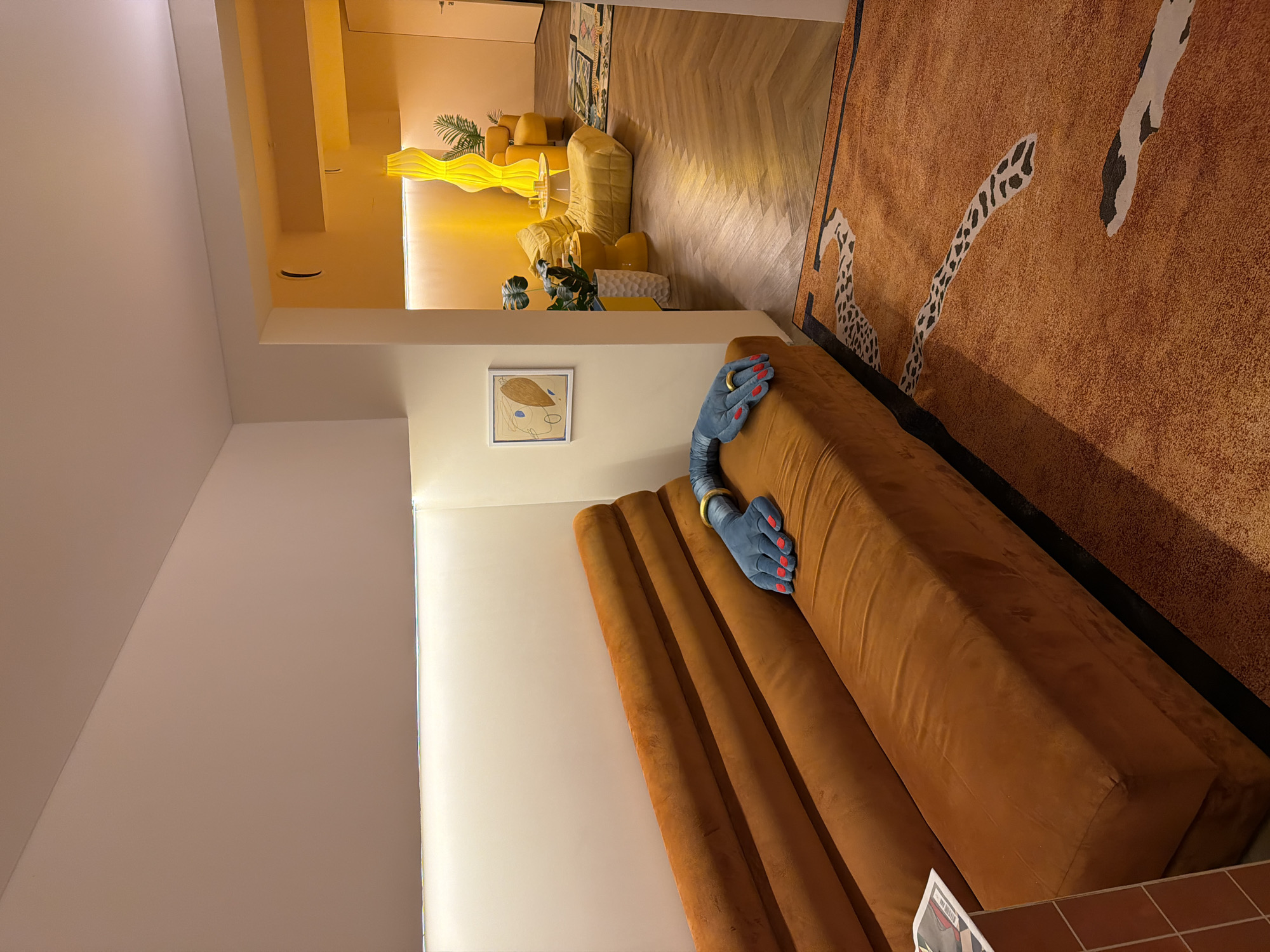 The image depicts a contemporary interior within an apartment in Moscow, Russia, designed by Nido Interiors. The scene is bisected into two distinct color-themed areas.
In the foreground and left midground, an elongated, ribbed sofa upholstered in burnt orange velour occupies the space. Resting on the right end of this sofa is a stylized blue sculpture resembling a human hand, complete with red painted fingernails and a gold bracelet on the wrist. Above the sofa, a linear white LED light strip illuminates a pale cream wall. The floor in this area is covered by a rectangular rug, predominantly orange, featuring abstract black and white organic patterns. A small section of a burnt orange tiled vertical surface is visible on the far left.
In the midground, a framed abstract artwork, depicting shapes in brown, blue, and white, hangs on a cream wall. This wall acts as a partition, delineating the burnt orange area from an adjoining room.
Through an open doorway on the right, a second room is visible, saturated in a warm, intense yellow light. This room contains multiple upholstered seating elements, including low chairs and cushion-like forms, all in varying shades of yellow. A tall, wavy yellow floor lamp stands prominently near the center. A small, round white side table, holding several clear drinking glasses, is situated among the yellow seating. Potted plants with large green leaves are placed throughout this yellow-themed area. The floor in this room features a chevron-patterned wooden parquet.
No individuals are present within the depicted scene.