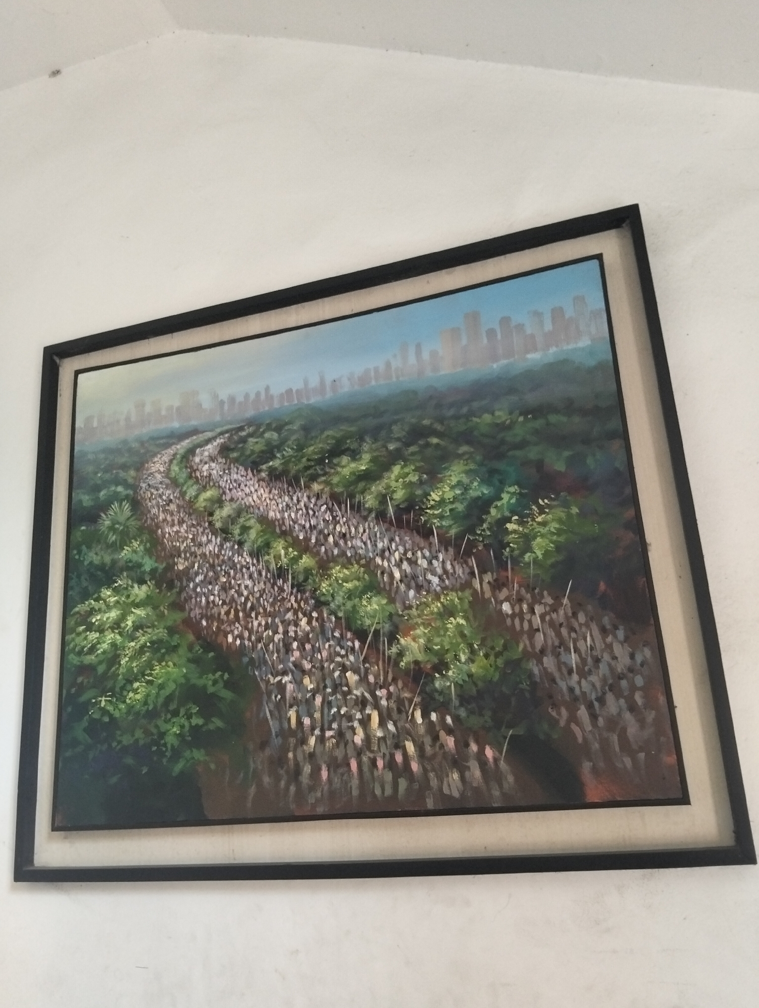 The image shows a framed painting hanging on a white wall, possibly in a domestic setting. The painting depicts a large crowd of people, seemingly a procession or march, winding through a lush, green landscape towards a cityscape in the background. The cityscape appears to be a distant skyline with numerous tall buildings. The painting style suggests an impressionistic or somewhat abstract representation of the scene, with visible brushstrokes and generalized figures. It is difficult to ascertain the exact location in Dhaka, Bangladesh, or even if the painting specifically depicts Dhaka. The scene evokes a sense of mass movement and possibly a political demonstration or religious gathering.