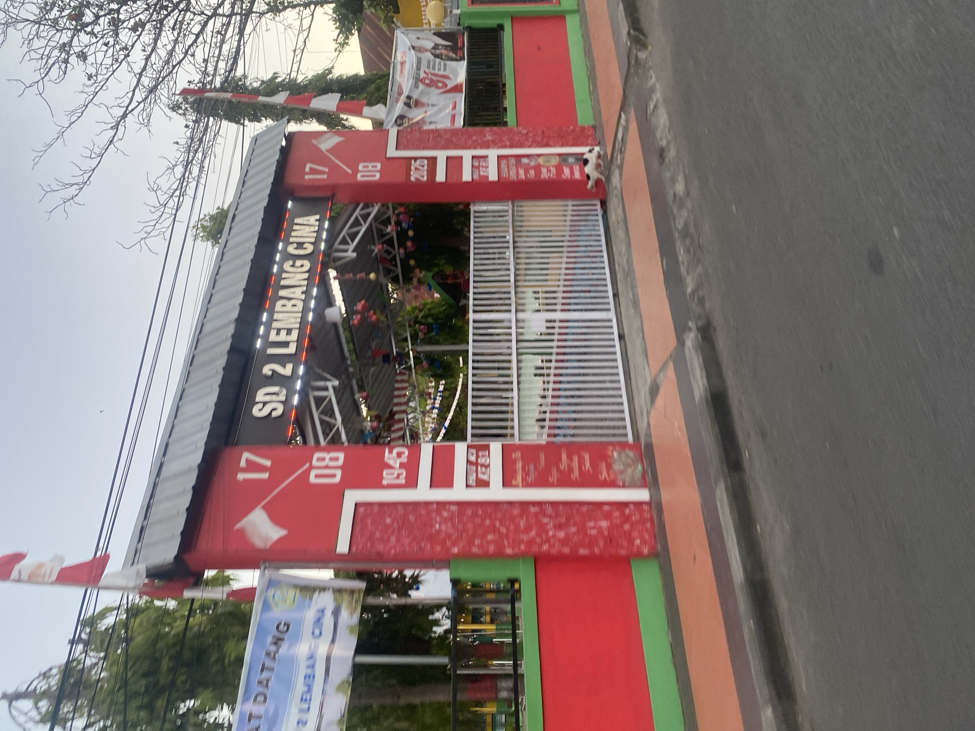 The image shows the entrance to SD 2 Lembang Cina, a school located in Bantaeng, Indonesia. The entrance is a large red gate with white and red flags. Above the gate, the school's name, "SD 2 LEMBANG CINA," is illuminated by white lights. To the left of the gate, a banner reads "SELAMAT DATANG" (Welcome) and the school's name in Indonesian. The gate has a white metal fence that leads into the school grounds.
On the right side of the entrance, a banner features the number "81" and portraits of two men, suggesting a celebration or event. Red and white decorations, possibly balloons and streamers, are visible inside the school grounds. A small black and white cat is walking near the base of the gate. The scene is a street view with a sidewalk in front of the school and a road in the foreground. The sky appears to be overcast.