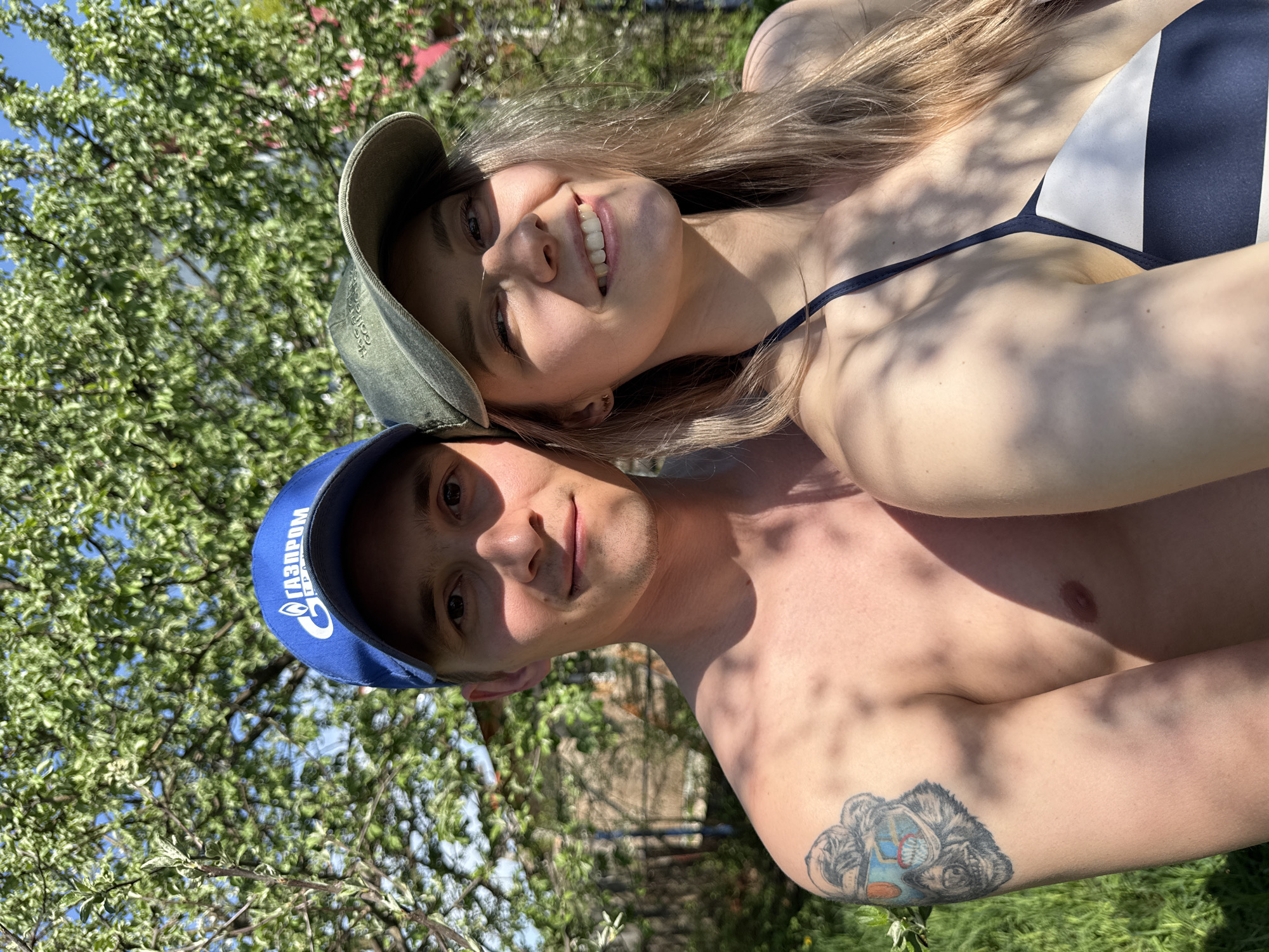 A man and woman are photographed outdoors in Chaikovsky, Russia. The man is shirtless and wears a blue baseball cap with a logo in white Cyrillic letters, and has a tattoo on his upper left arm. The woman wears a green baseball cap, and appears to be wearing a black and white swimsuit top. The photo is taken under a tree.