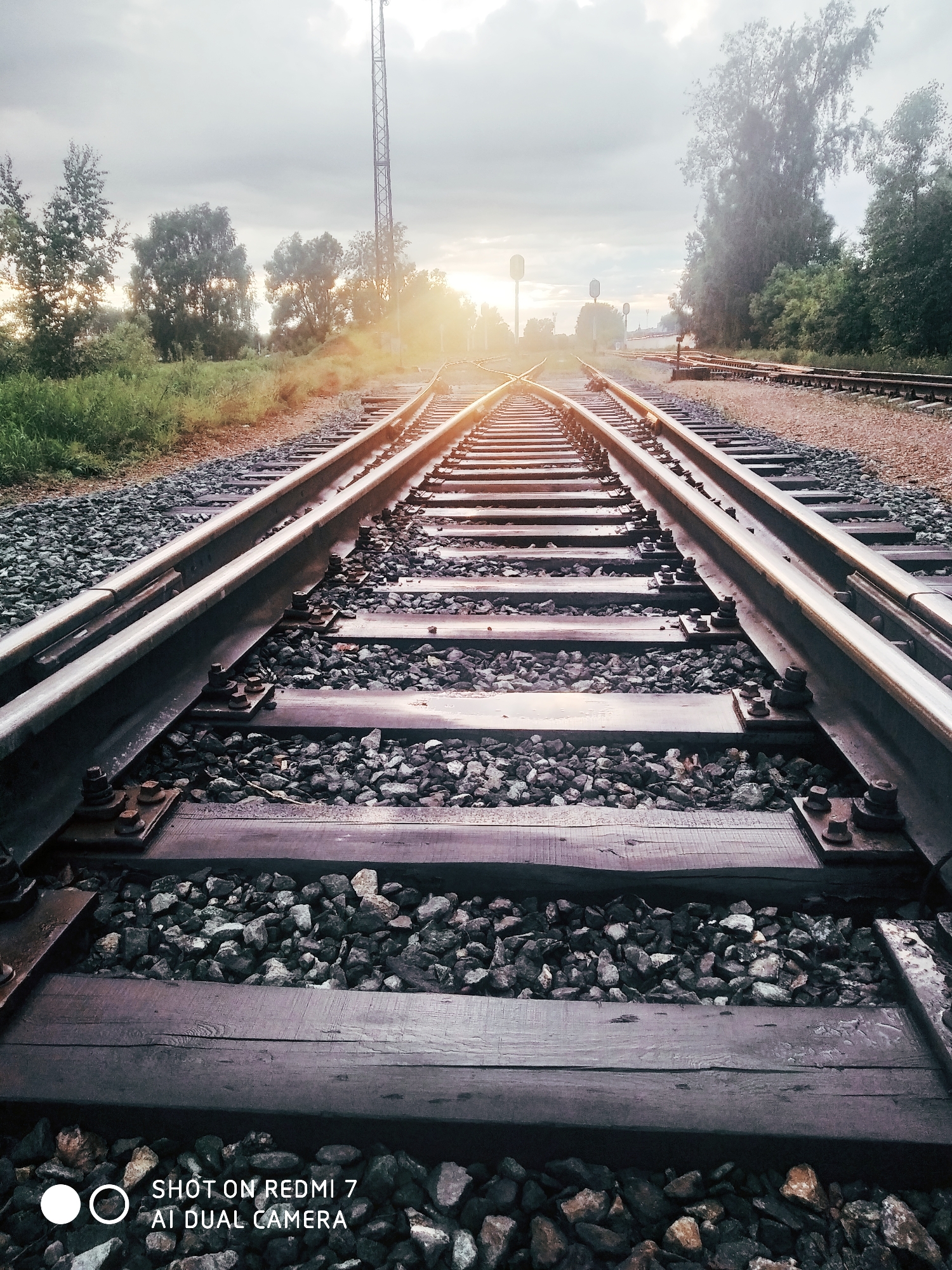 A low-angle photograph captures a railway track scene at either sunrise or sunset, characterized by a prominent sun flare. The image focuses on two main railway tracks, constructed from dark metal rails secured to weathered wooden sleepers (ties) that are set within a bed of gray and brown crushed stone ballast. The tracks extend from the immediate foreground, where details of the fastenings and ballast are clearly visible, into the hazy distance, converging and branching into multiple lines in the middle ground, indicating a railway switch or turnout.
A strong, warm lens flare emanates from the low-lying sun on the right side of the frame, casting a golden glow across the tracks and contributing to a soft, atmospheric haze. On both sides of the tracks, green grass and wild vegetation grow alongside a dense line of trees with varied foliage, bordering the railway line. A tall, slender lattice mast, possibly a signal or transmission tower, is visible on the left side of the tracks, extending towards the cloudy sky. Further along the tracks in the distance, subtle details of other railway infrastructure, such as signals, can be discerned. The sky is largely overcast with diffused light, yet the low sun creates a dramatic highlight.
No people are present in the image, emphasizing the quiet solitude of the railway environment. A watermark in the bottom left corner reads: "SHOT ON REDMI 7 AI DUAL CAMERA".